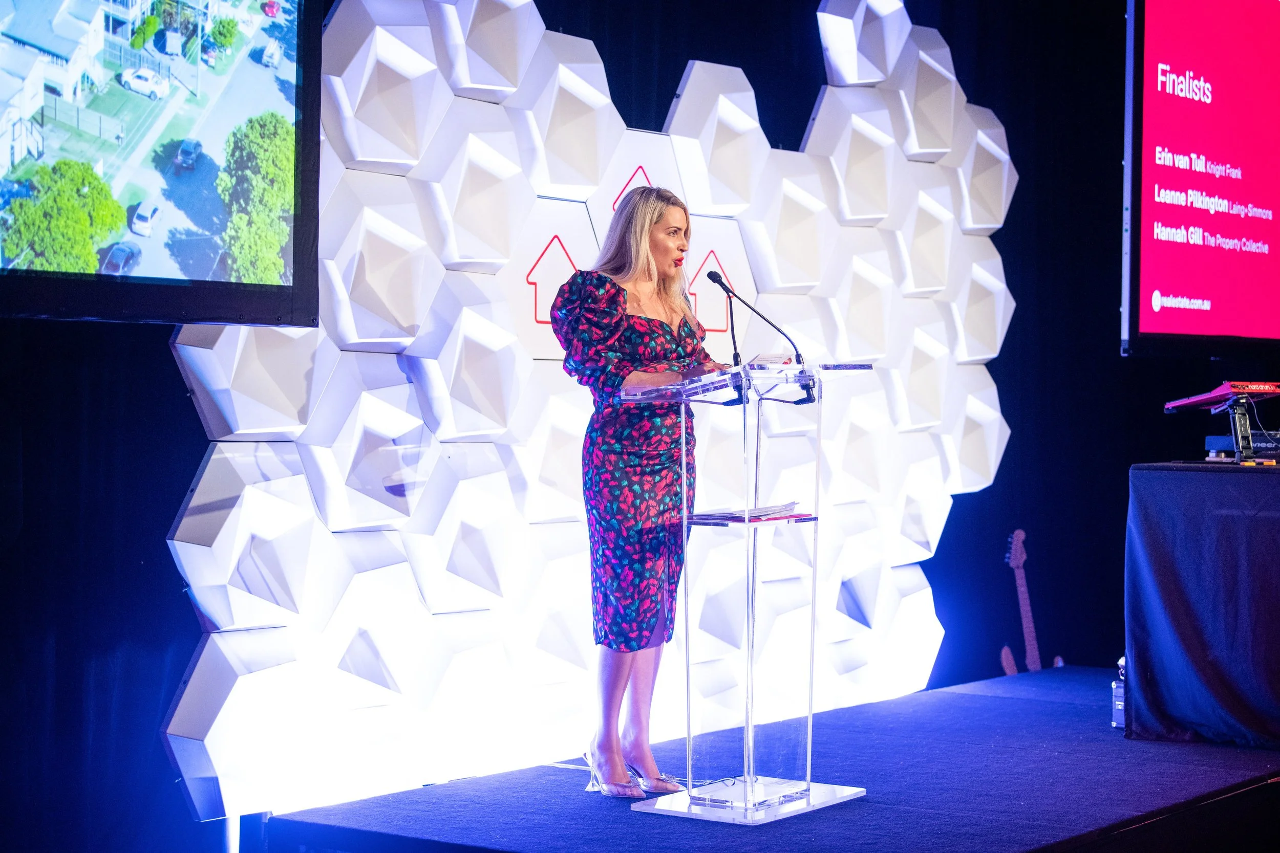 A woman with blonde hair wearing a colorful floral dress stands at a clear podium giving a presentation at an event. Behind her is a decorative white wall and two large screens, one displaying aerial view of a neighborhood and the other a list of fin