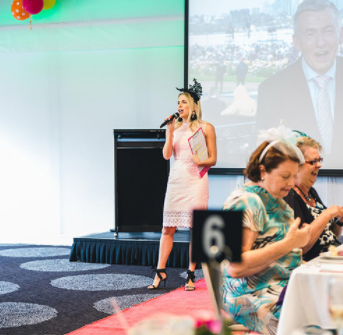 Young woman in pink dress speaking into a microphone at a banquet, with elderly women seated at a table in the foreground, and a large screen behind her displaying a man in a suit.