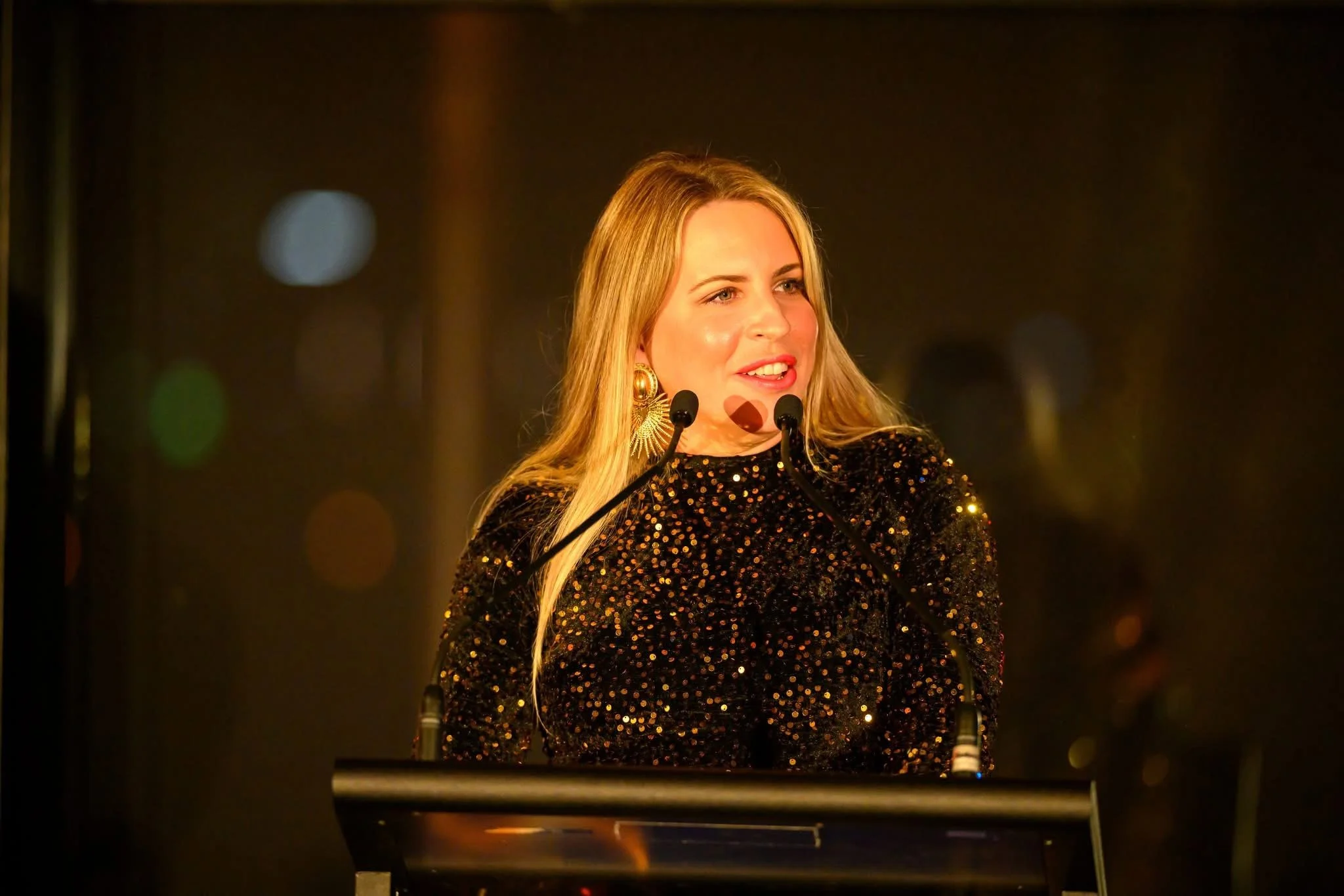 A woman with long blonde hair wearing a black, sequined dress speaking at a podium with two microphones in front of a dark, blurred background with colorful bokeh lights.