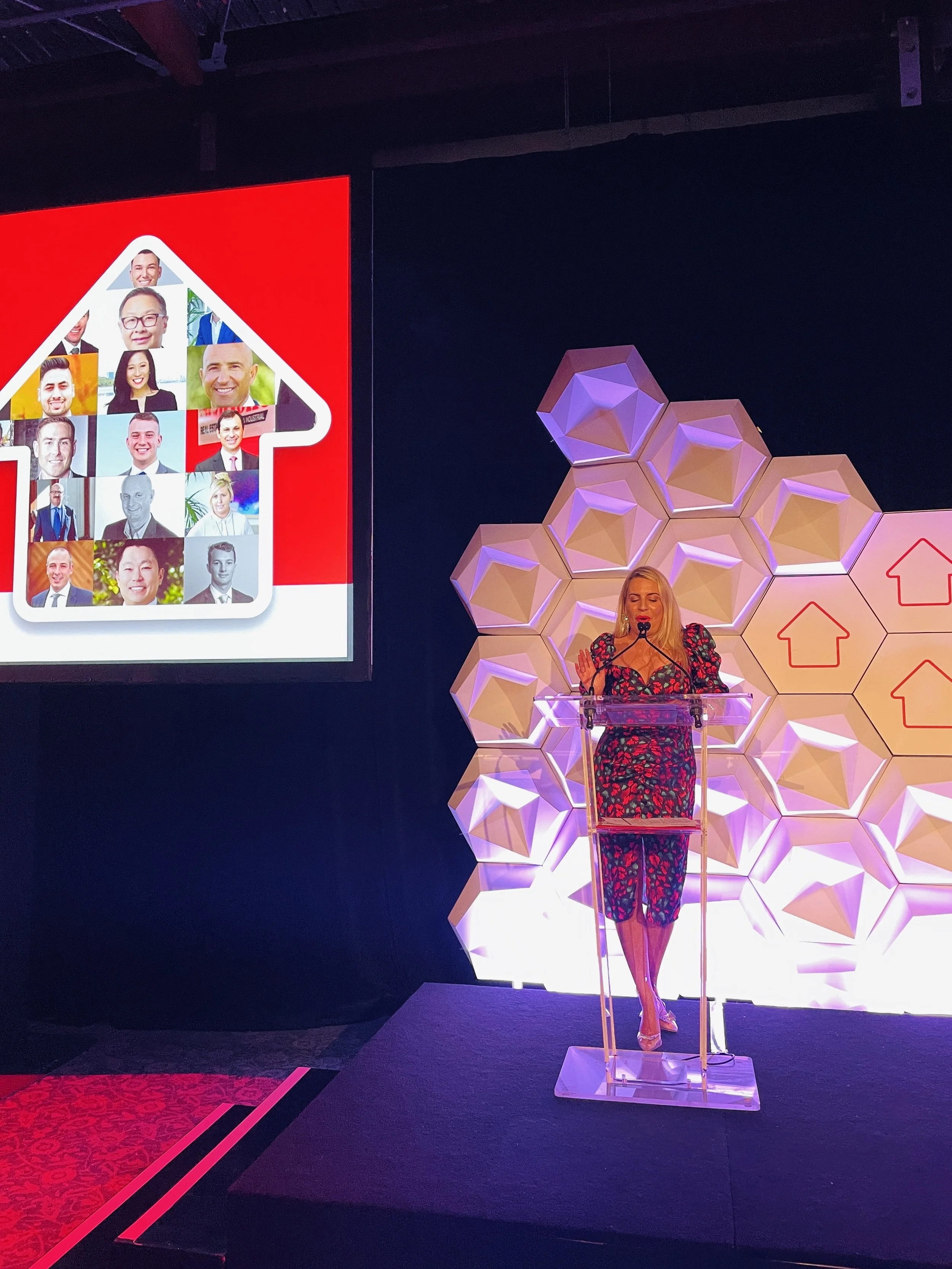 A woman giving a presentation at a conference, standing behind a clear podium with a microphone, with a geometric honeycomb-patterned backdrop and a large screen displaying an infographic of a house filled with photos of diverse people.
