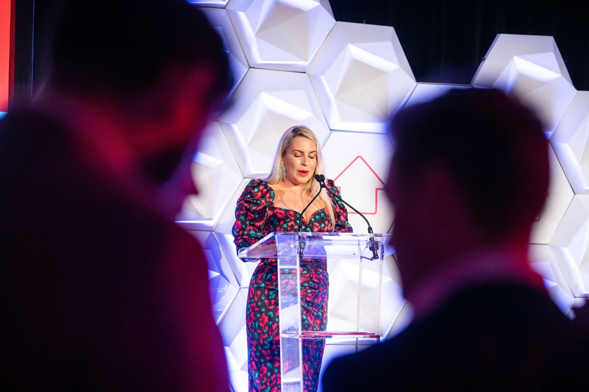 A woman in a colorful dress speaking at a podium during a formal event, with a geometric backdrop and audience visible in the foreground.