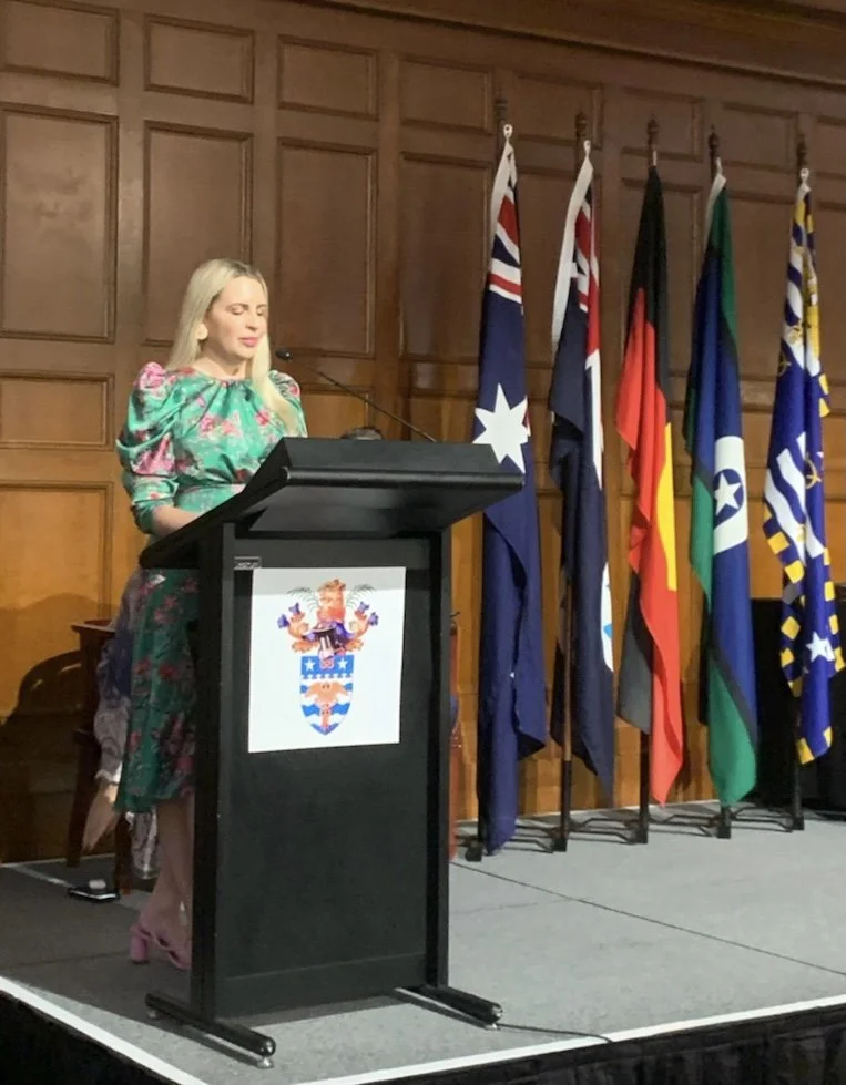 A woman with blonde hair in a colorful dress stands at a podium with a coat of arms, speaking or making a speech. Behind her, seven flags are displayed, representing Australia, New Zealand, the United Kingdom, Germany, Italy, Greece, and another coun