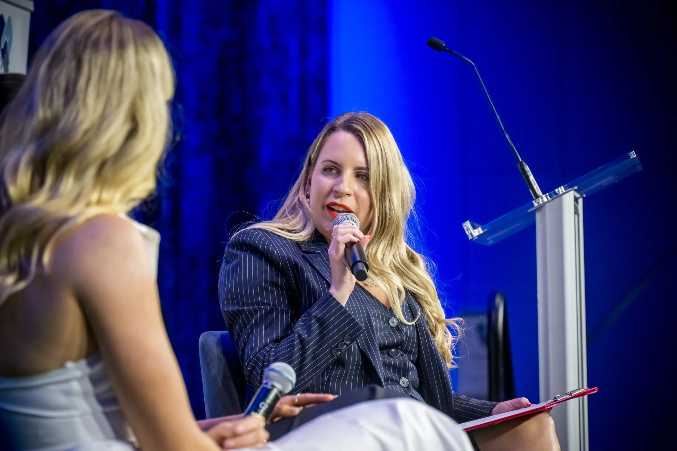 Two women participating in a panel discussion, with one woman speaking into a microphone and the other woman listening.