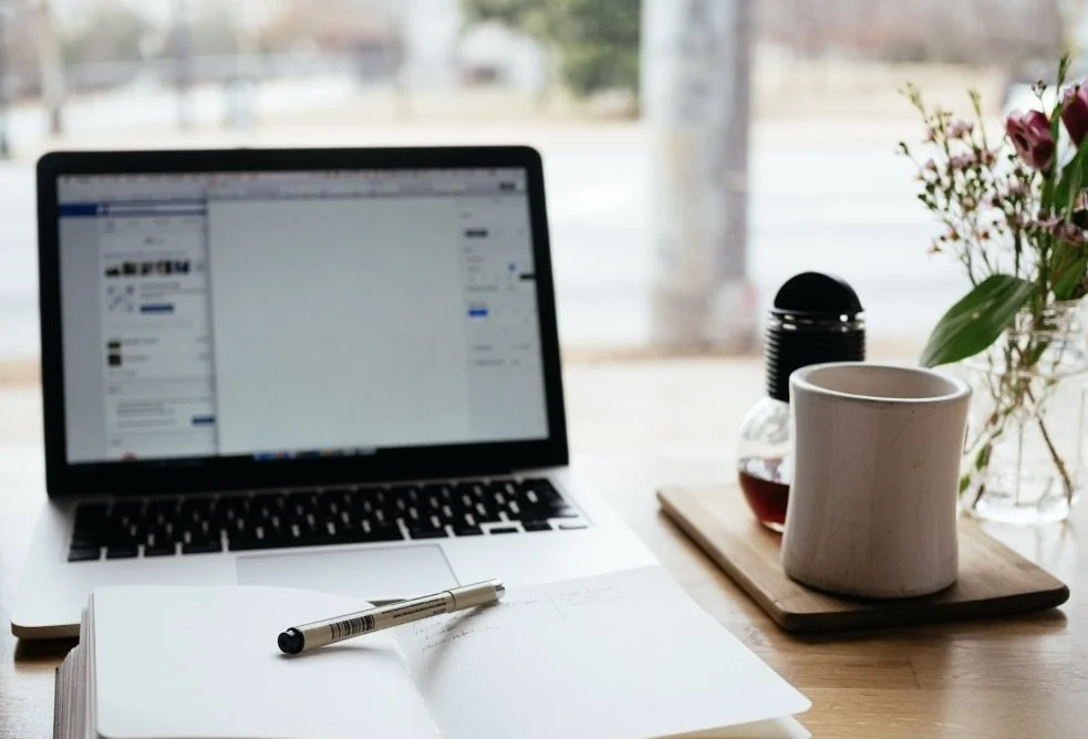 Laptop on a desk with an open notebook, pen, coffee cup, and flowers.
