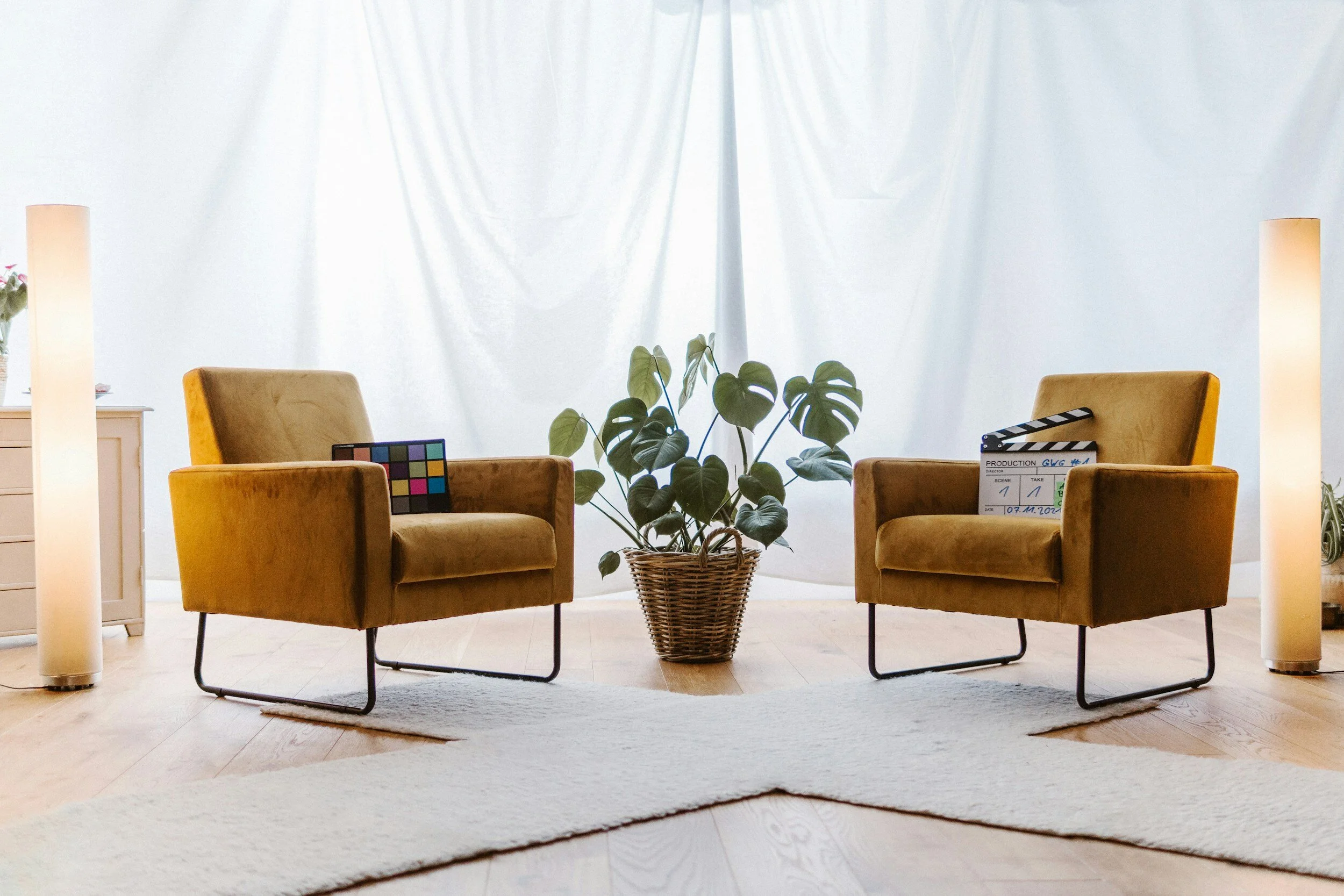 Two mustard yellow armchairs face each other with a wicker basket containing a monstera plant between them. A soft white rug is on the wooden floor. A tall lamp is beside each chair, and a white background curtain is visible. A film clapboard rests on one chair, and a color chart on the other.