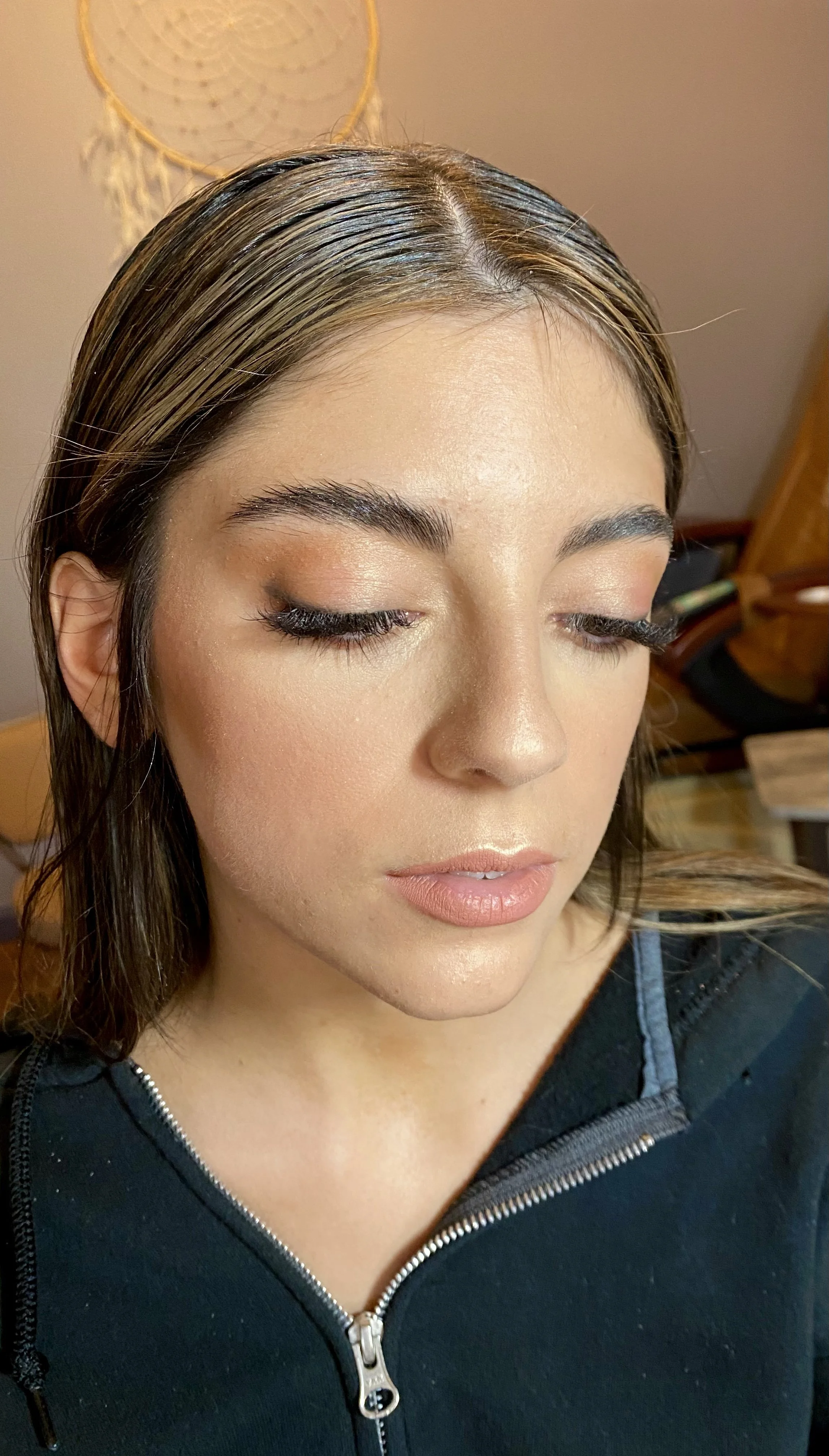 Close-up of a woman with closed eyes showcasing makeup, wearing a black zip-up hoodie, with a dreamcatcher hanging on the wall in the background.