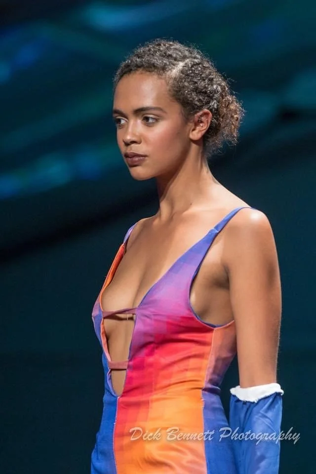 A woman with curly hair wearing a colorful dress with cutout details, standing against a dark background.