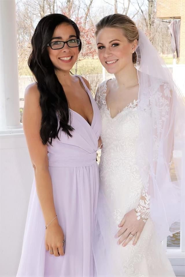 Two women standing together, one in a lavender dress and the other in a white wedding gown with a veil, smiling.