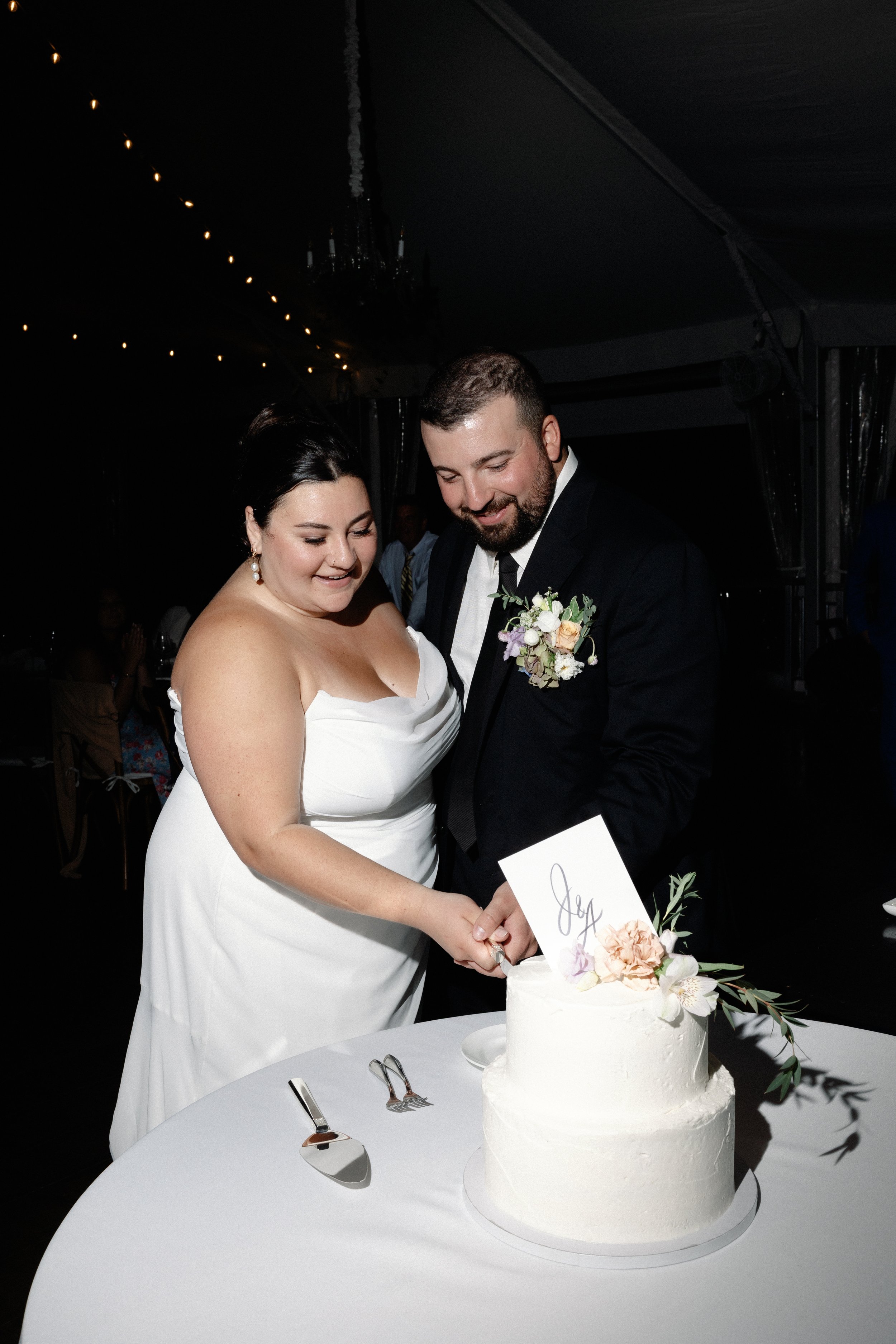 A bride and groom cutting a wedding cake at their reception, with a dark background and string lights overhead.