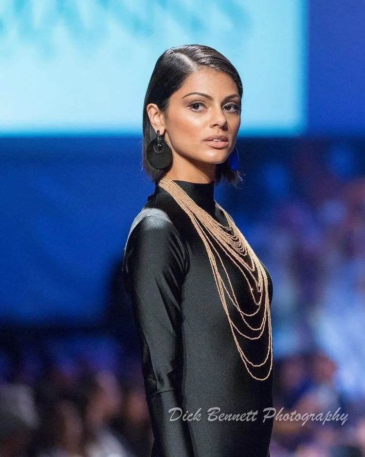 A woman with short black hair wearing large black earrings and layered necklaces, dressed in a black satin dress, standing on a runway or stage with an audience in the background.