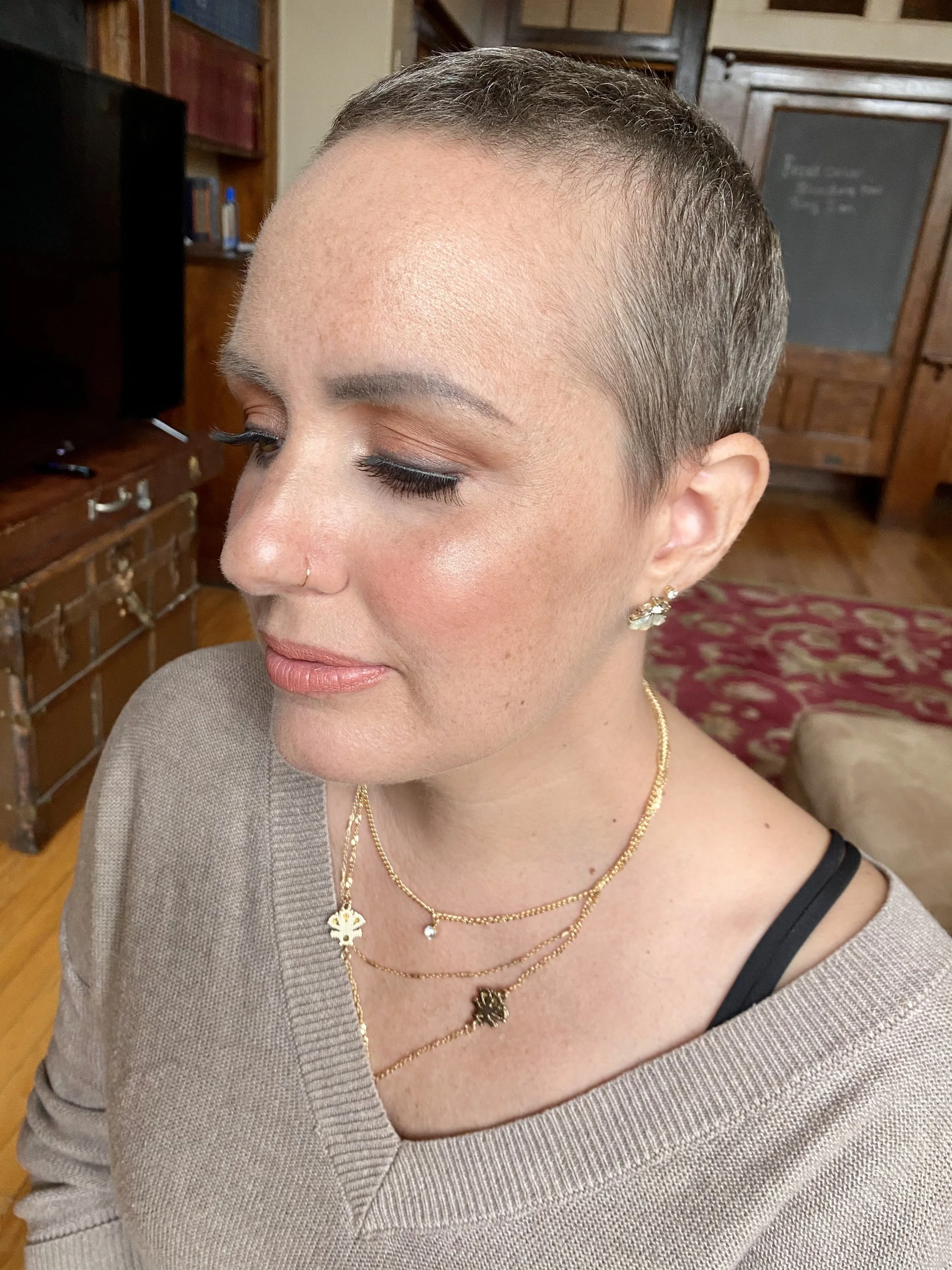 Close-up of a woman with a short buzzed haircut, wearing makeup, gold jewelry, and a beige sweater, inside a room with wooden furniture.