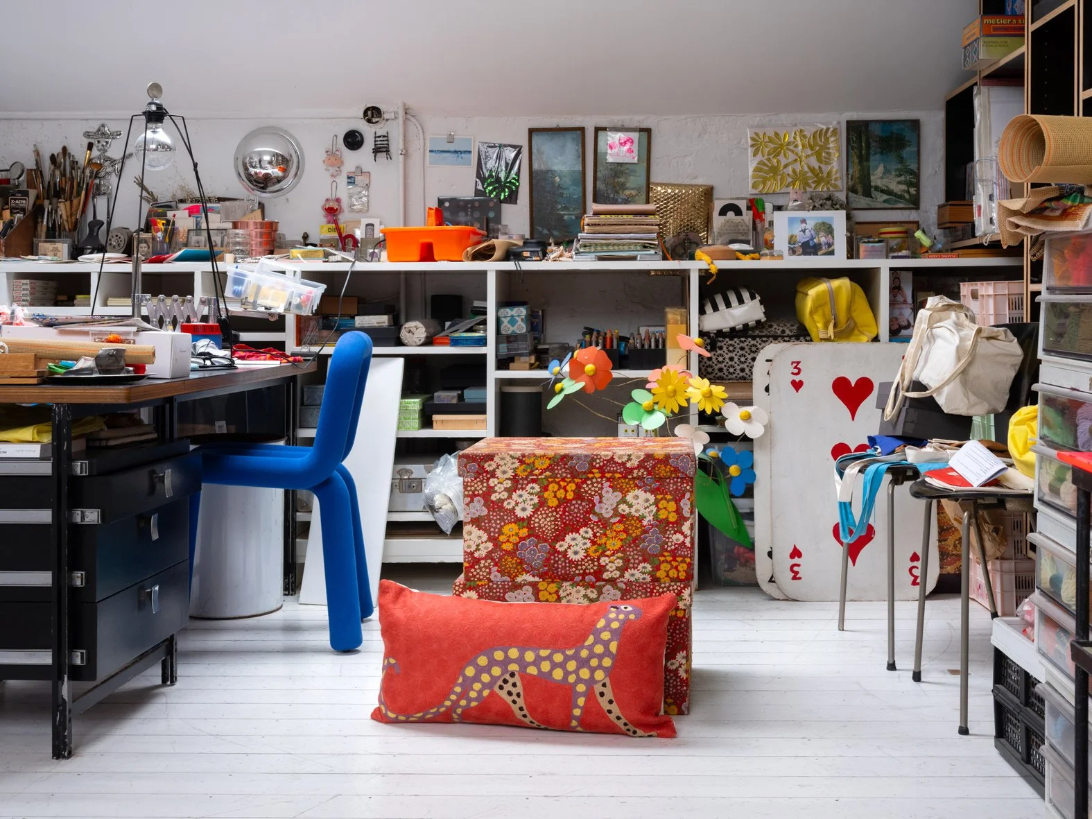 Un atelier rempli d'objets divers, comprenant une chaise bleue, un bureau avec des outils, des boîtes de rangement, des cadres accrochés au mur, un grand paquet cadeau orné de fleurs, un coussin avec un lézard stylisé, et d'autres articles d'art ou d
