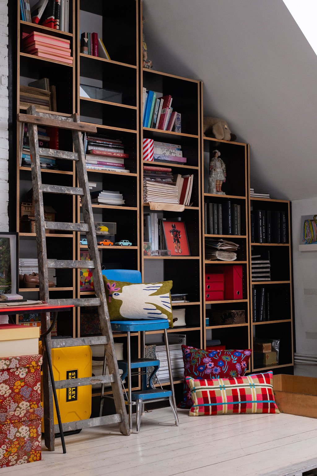 Une grande étagère en bois remplie de livres, objets décoratifs et magazines, située dans une pièce avec un plafond en pente, avec un escabeau en bois ancien, des coussins colorés et une chaise bleue.