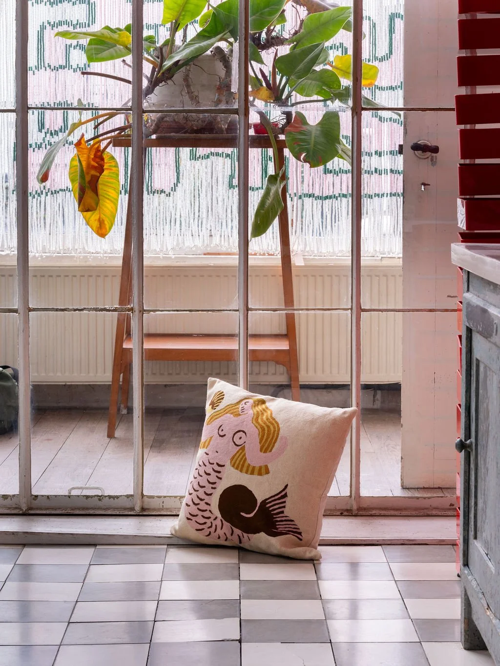 Coussin avec une illustration de sirène et de poisson sur le sol en carreaux devant une porte en verre avec des plantes vertes en arrière-plan