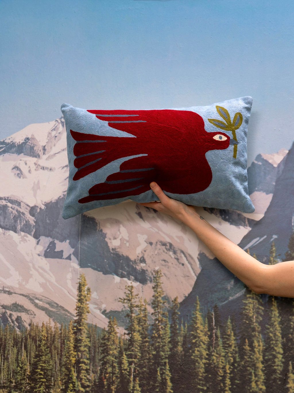 Un coussin rectangulaire avec un motif d'oiseau rouge tenant une branche verte, tenu par une main devant un mur avec une image de montagnes et de forêt.