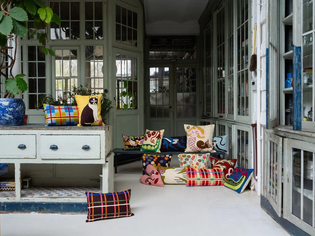Intérieur de véranda ou salon avec des coussins colorés et décoratifs, certains représentant des animaux ou avec des motifs variés, sur une vieille armoire et un sol en béton, avec une ambiance conviviale et artistique.