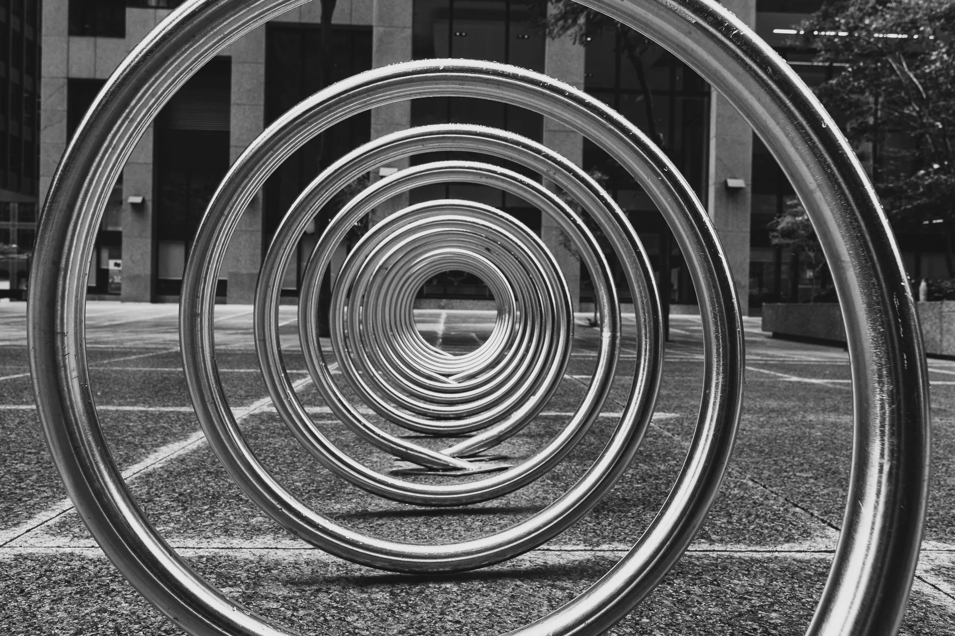 Black and white photo of concentric metal circles on a city sidewalk, with buildings and trees in the background.
