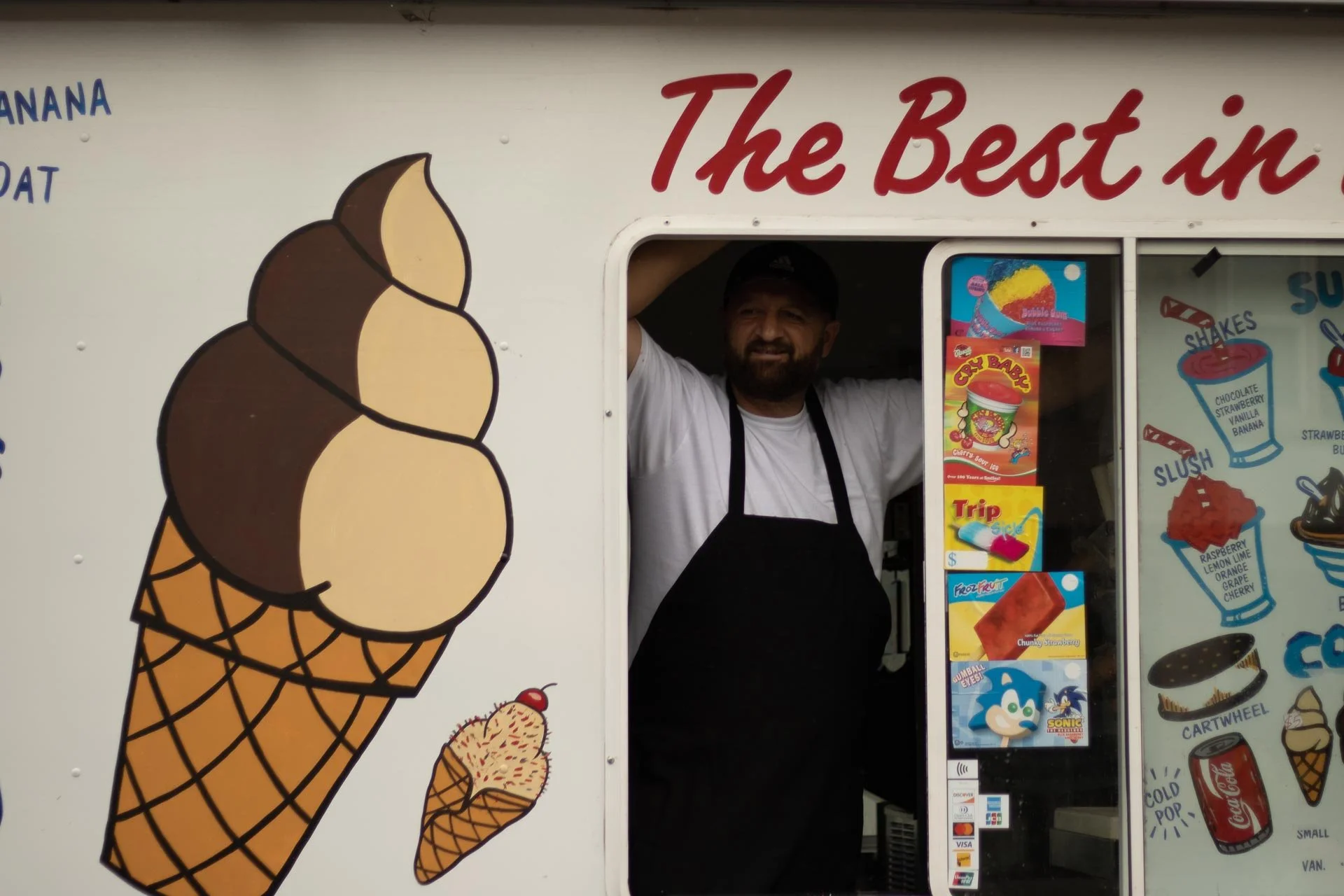 Ice cream vendor standing inside a colorful ice cream truck with images of ice cream cones, floats, and shakes on the side. The truck has the phrase 'The Best in' written on it.