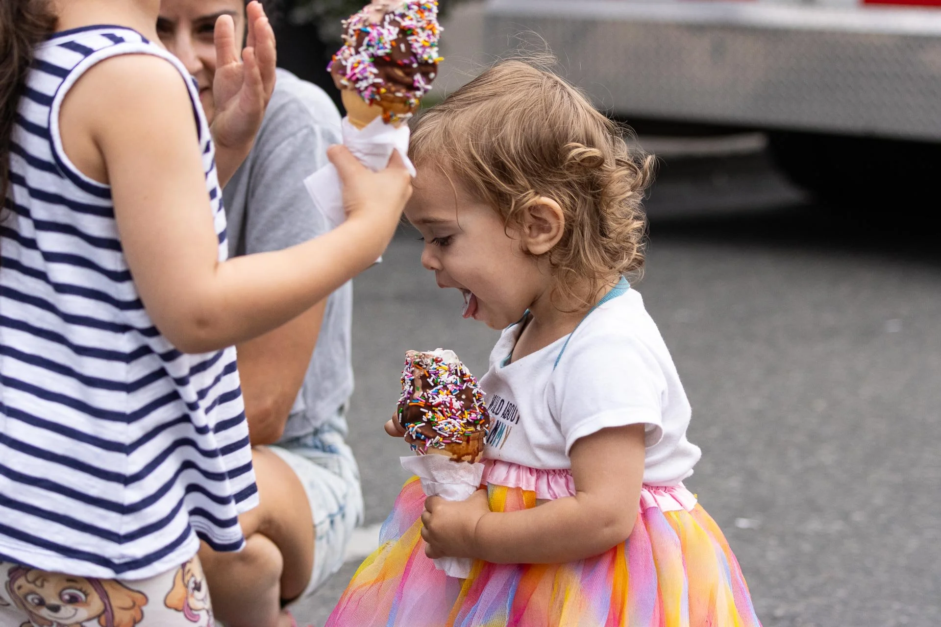 Child with curly hair wearing a colorful rainbow tutu dress, holding a chocolate ice cream cone with sprinkles, smiling with tongue out.