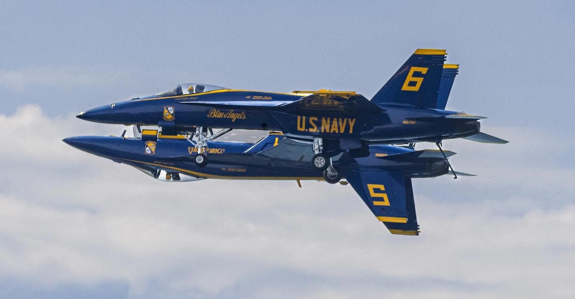 A United States Navy Blue Angels fighter jet performing a mirror flight, flying inverted above clouds, painted in blue and yellow with the tails numbered 5 and 6.