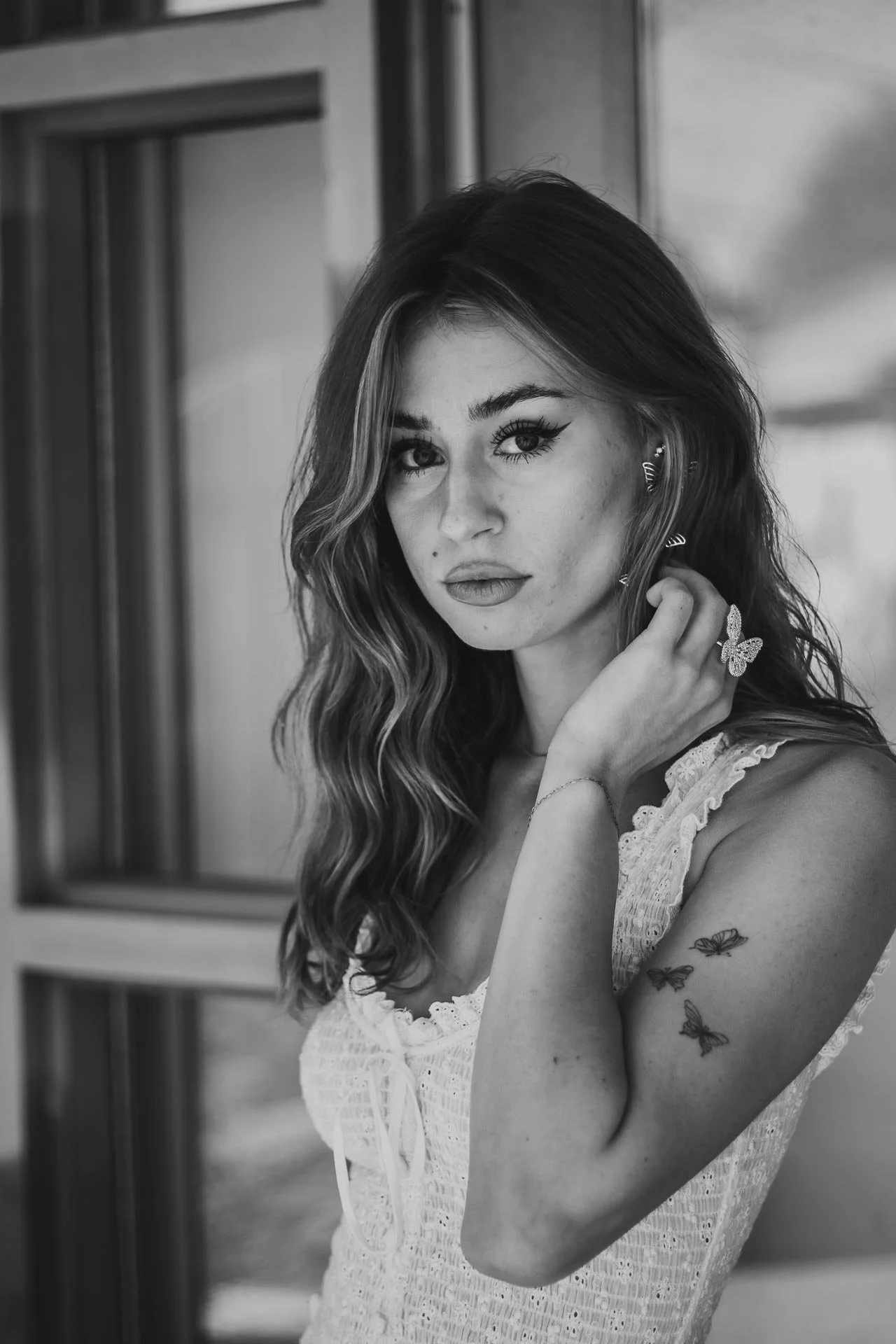 A black-and-white portrait of a young woman with long wavy hair, wearing earrings, a butterfly ring, and a sleeveless dress, standing near a window.