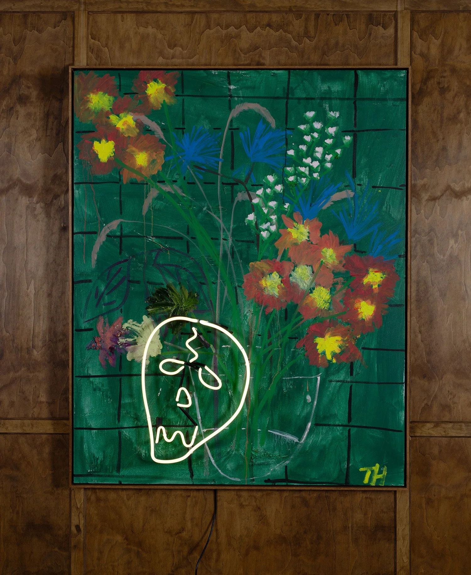 Colorful painting of flowers on a green background with a white neon skull outline and the signature 'TH' in yellow in the bottom right corner, mounted on a wooden wall.