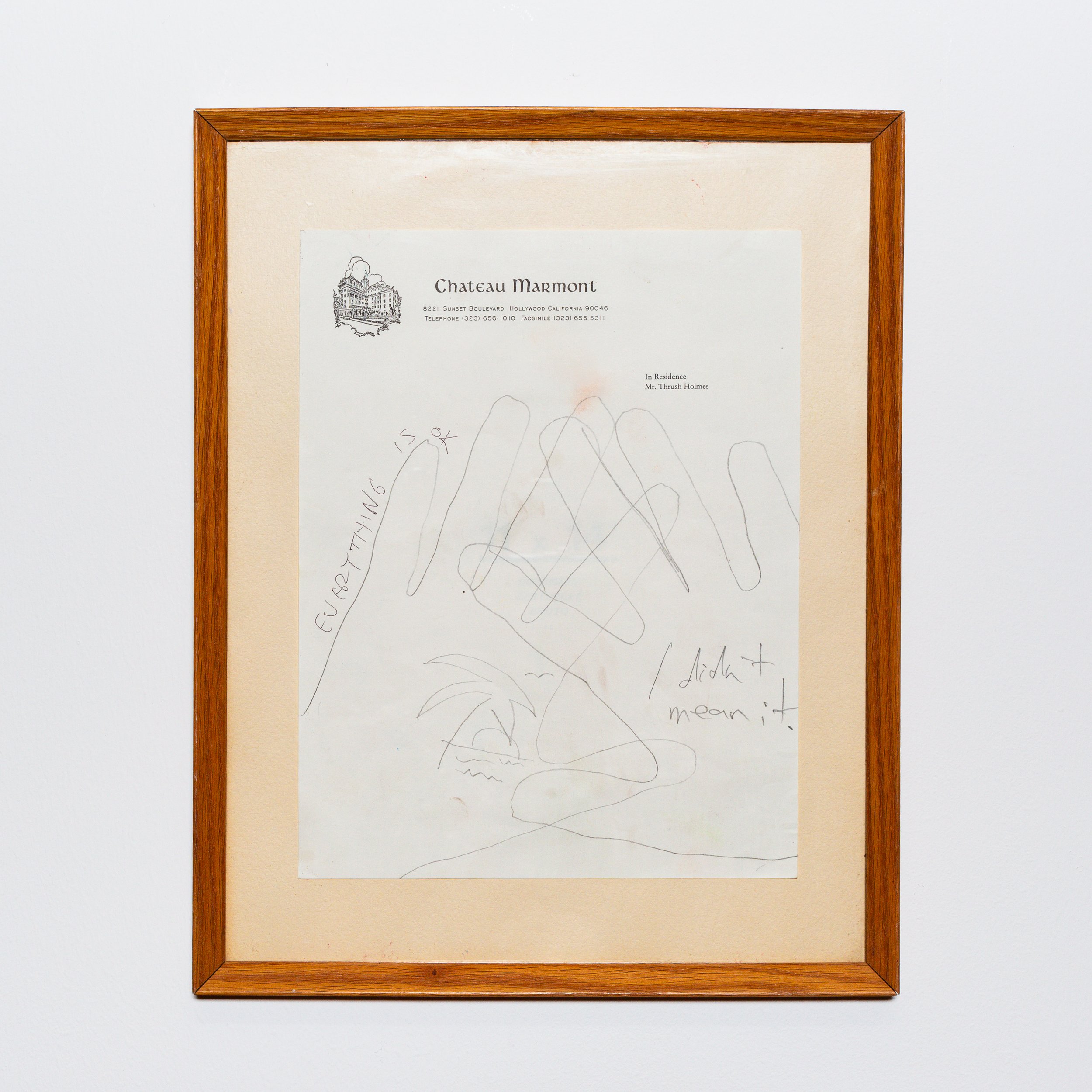A framed piece of paper with handwritten scribbles and some printed text, hanging on a plain white wall.