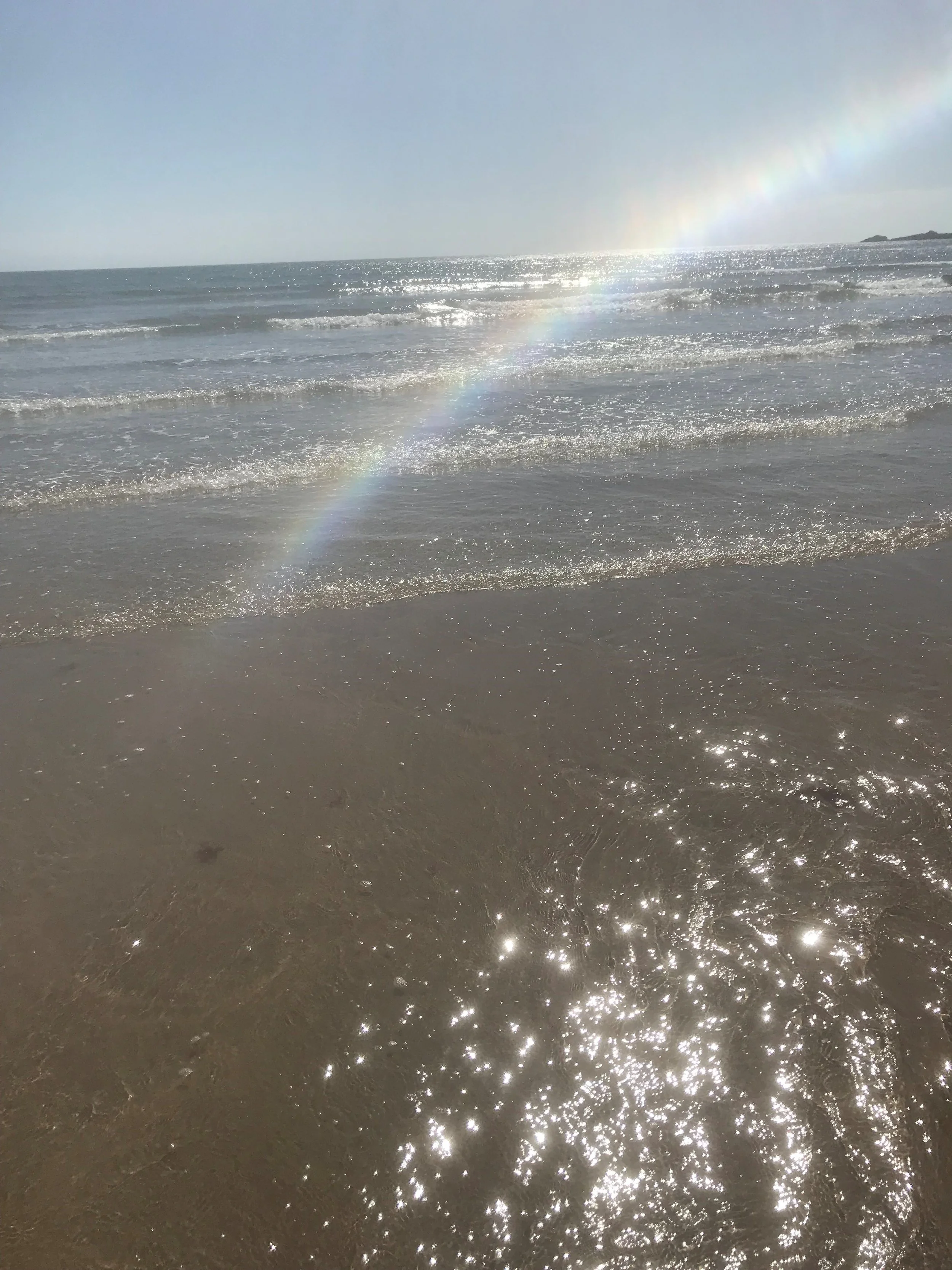 Ocean shoreline with gentle waves and a rainbow in the sky, sunlight reflecting off the water.