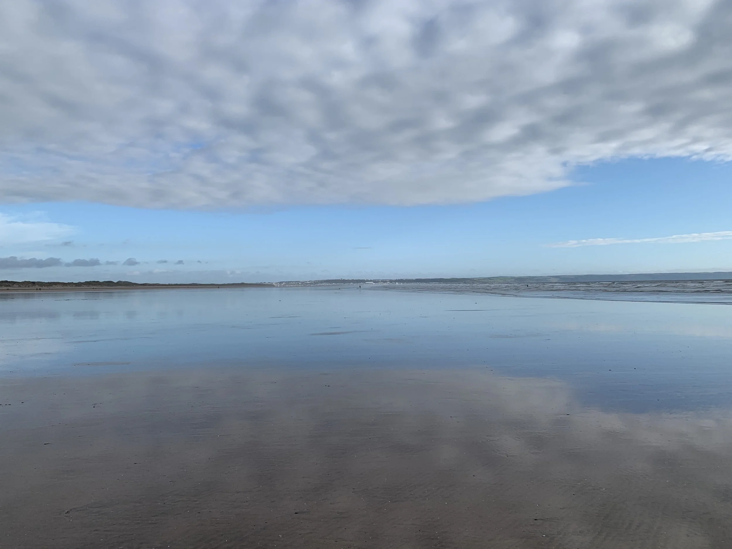 A calm beach with wet sand reflecting a blue sky with scattered clouds, distant shoreline on the horizon.