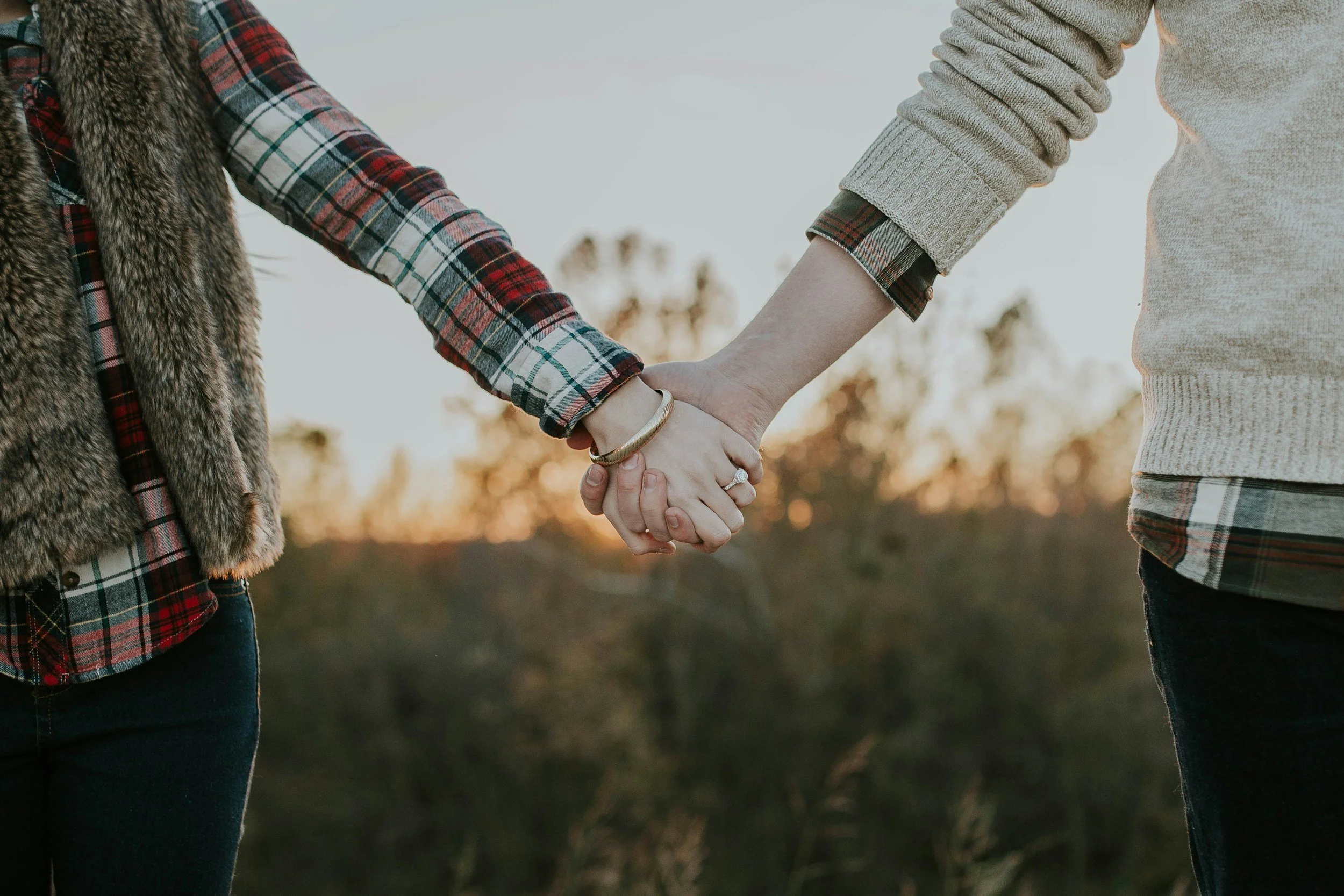 Two people holding hands outdoors during sunset, one wearing a plaid shirt and faux fur vest, the other in a sweater, with trees in the background.