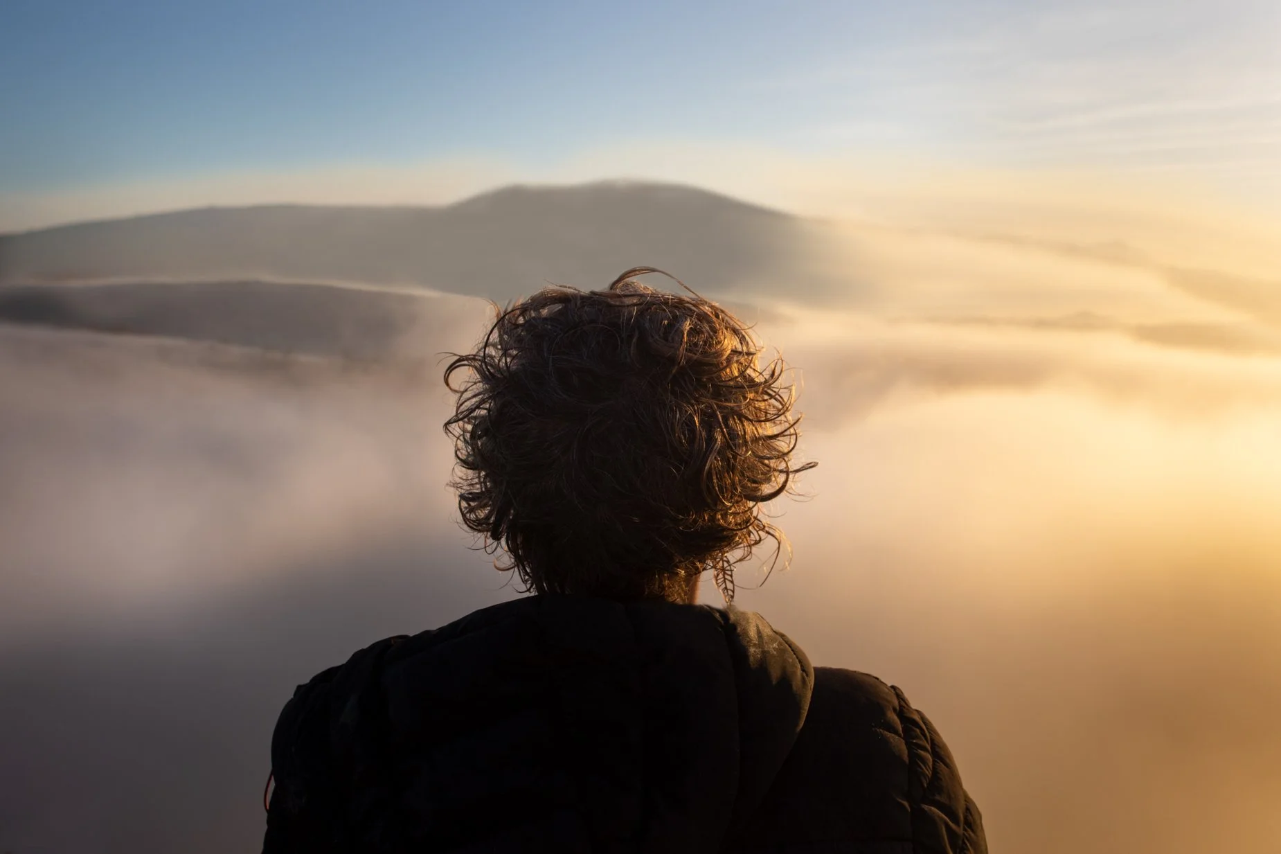 Person with curly hair wearing a green sweater watching the sunrise or sunset outdoors, with trees and clouds in the background.