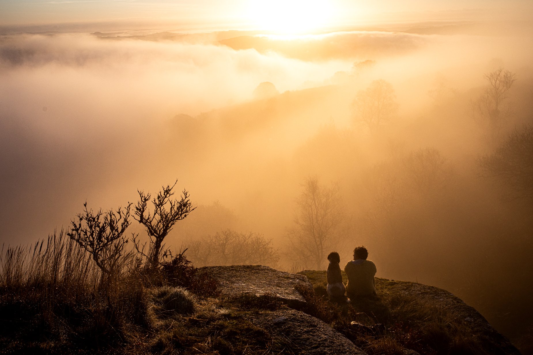 A person and a child sitting on a rocky terrain, watching a foggy landscape at sunset with trees and distant hills.