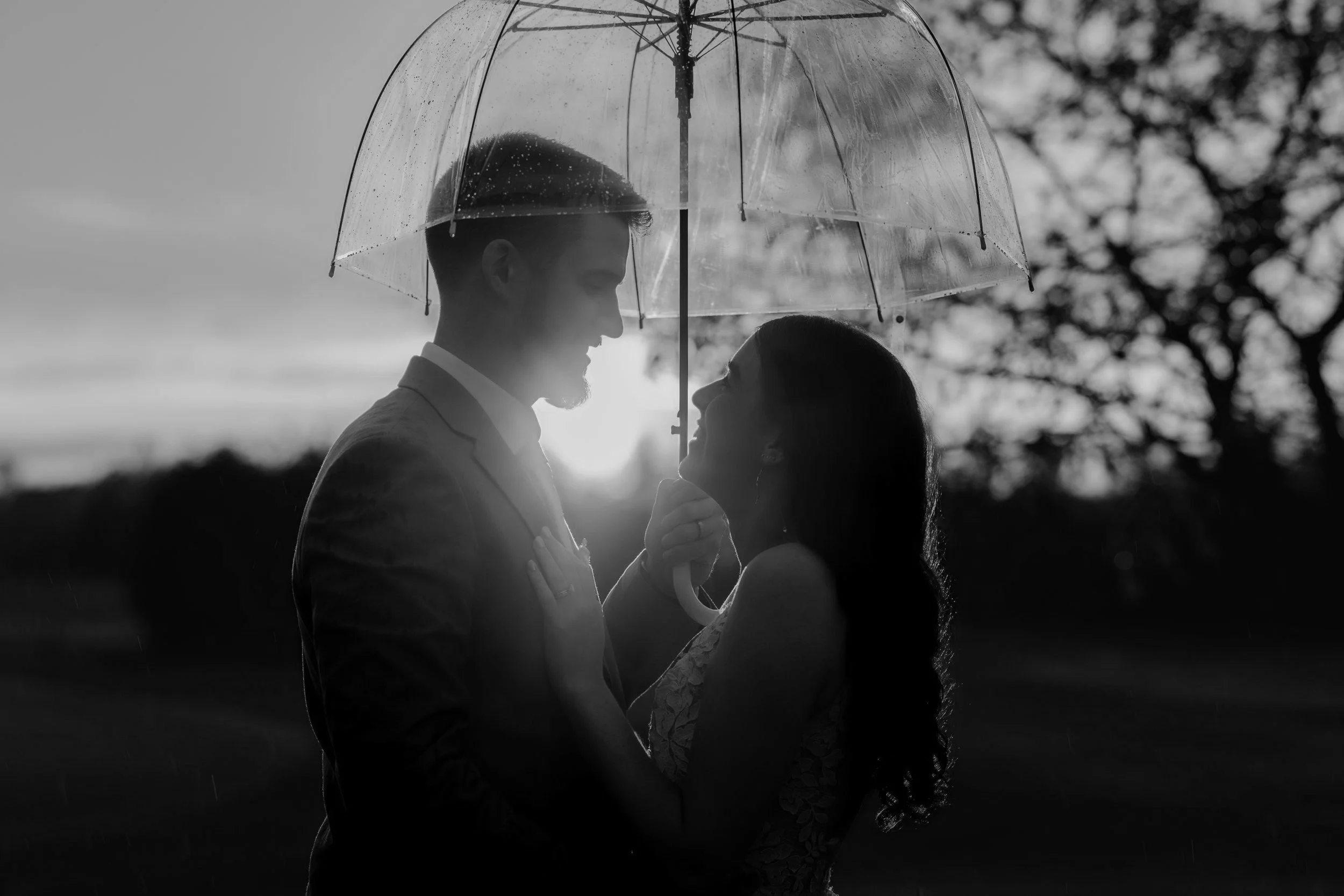 Silhouette of a couple under a transparent umbrella during rain at sunset, facing each other affectionately.