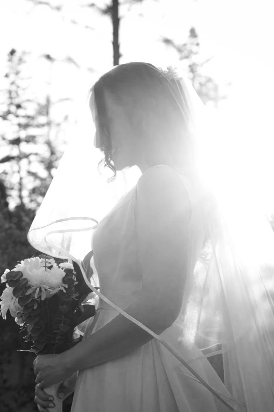 A black and white photo of a bride holding a bouquet of flowers, with sunlight shining behind her, creating a silhouette effect.