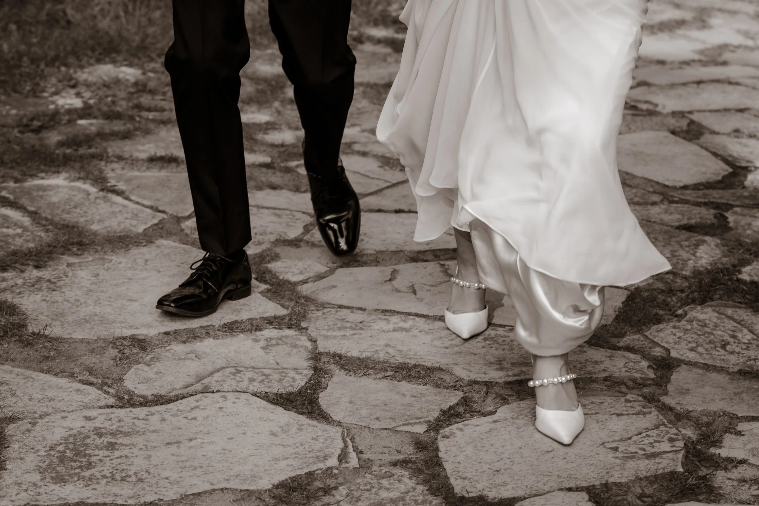 Close-up of a bride and groom's lower legs and feet walking on a stone pathway, with the bride wearing white high heels and pearl anklets, and the groom in black dress shoes and pants.