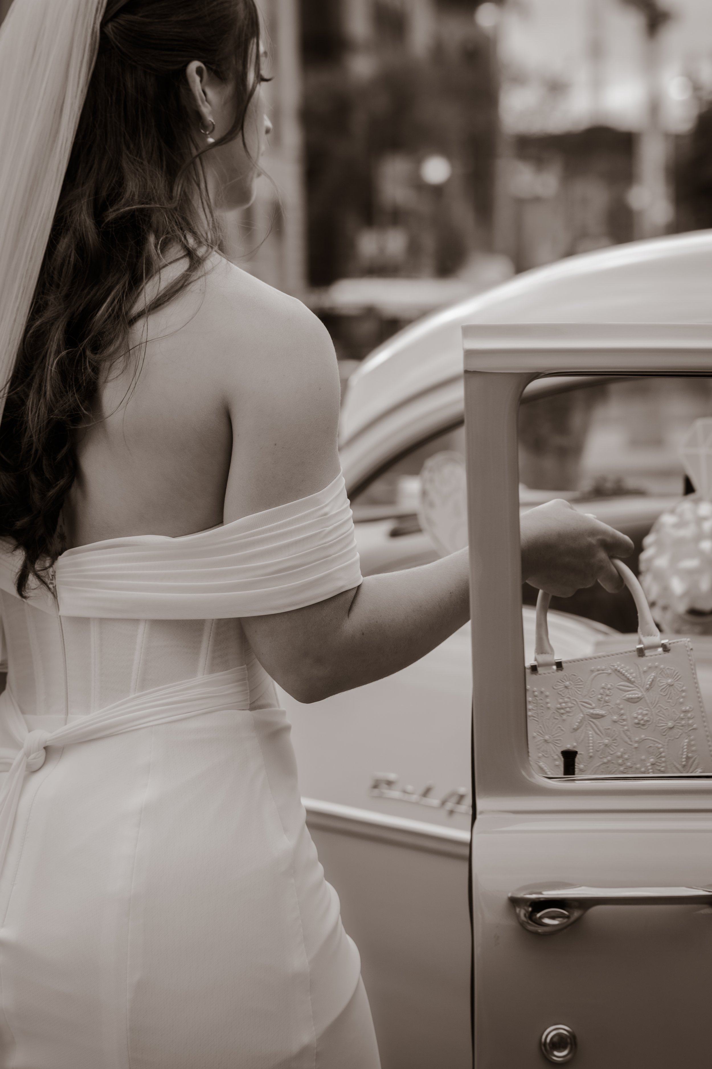 A woman in a wedding dress having her corset laced up by another person, with curly hair and detailed lace fabric on the dress.