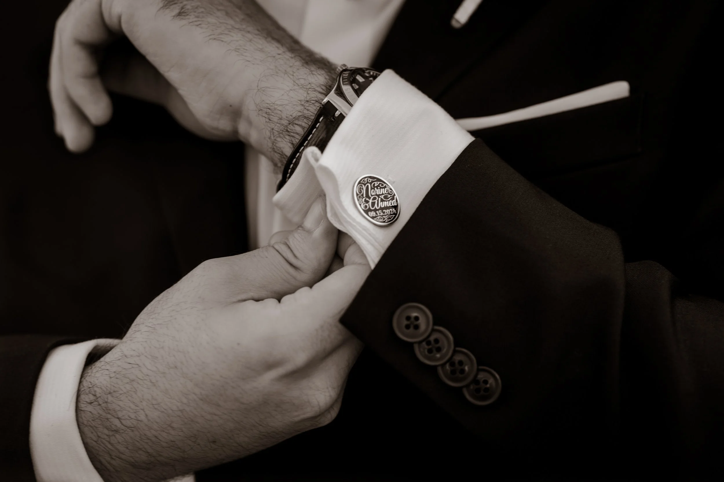 Close-up of a person adjusting their cufflinks on a tuxedo sleeve, with a wedding date badge on the cuff.