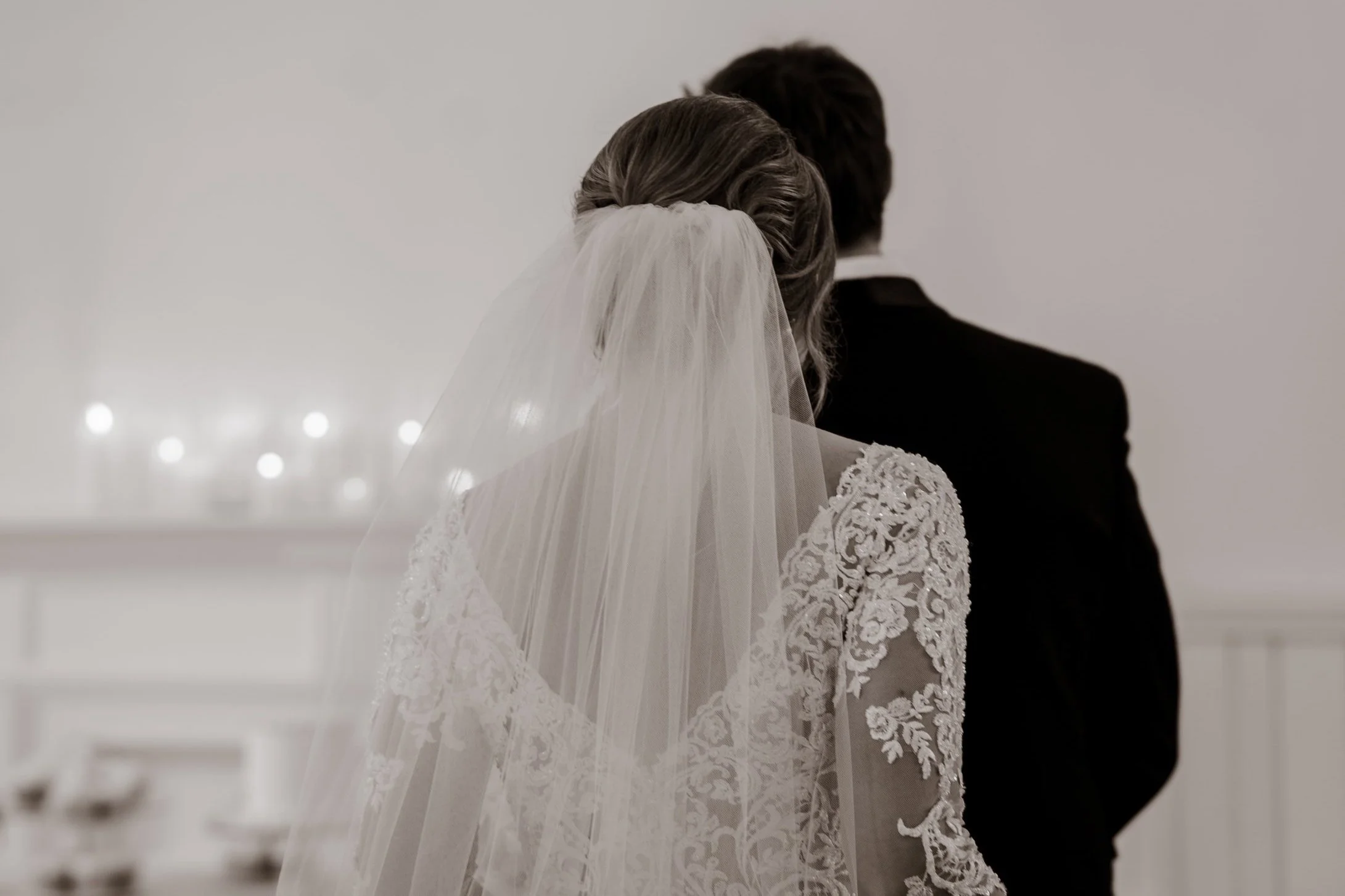 A bride and groom facing away during a wedding ceremony, The bride wears a lace wedding dress and veil, the groom a dark suit, in a minimalistic indoor setting.