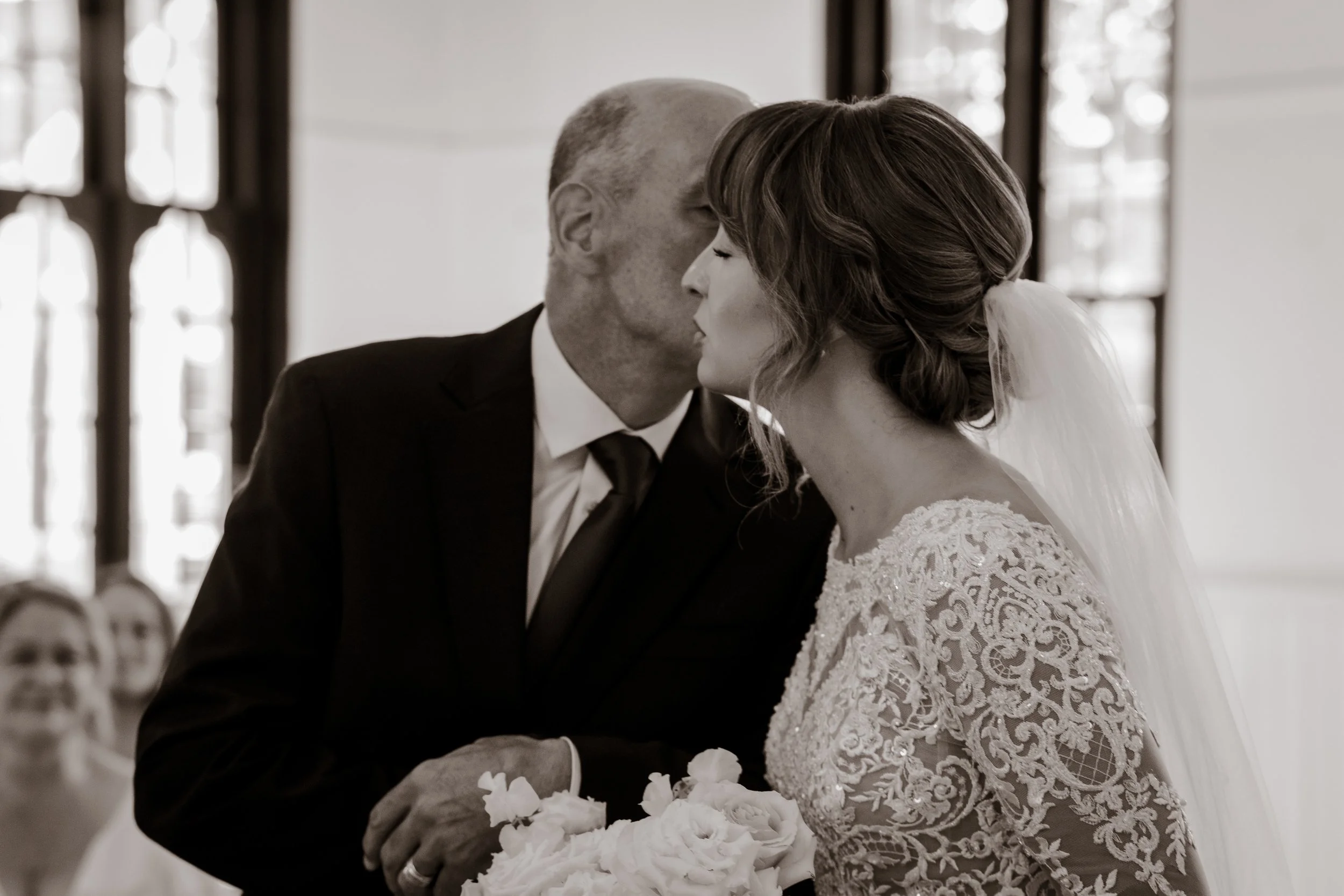 A bride and an older man, possibly her father, share a kiss during a wedding ceremony inside a church.
