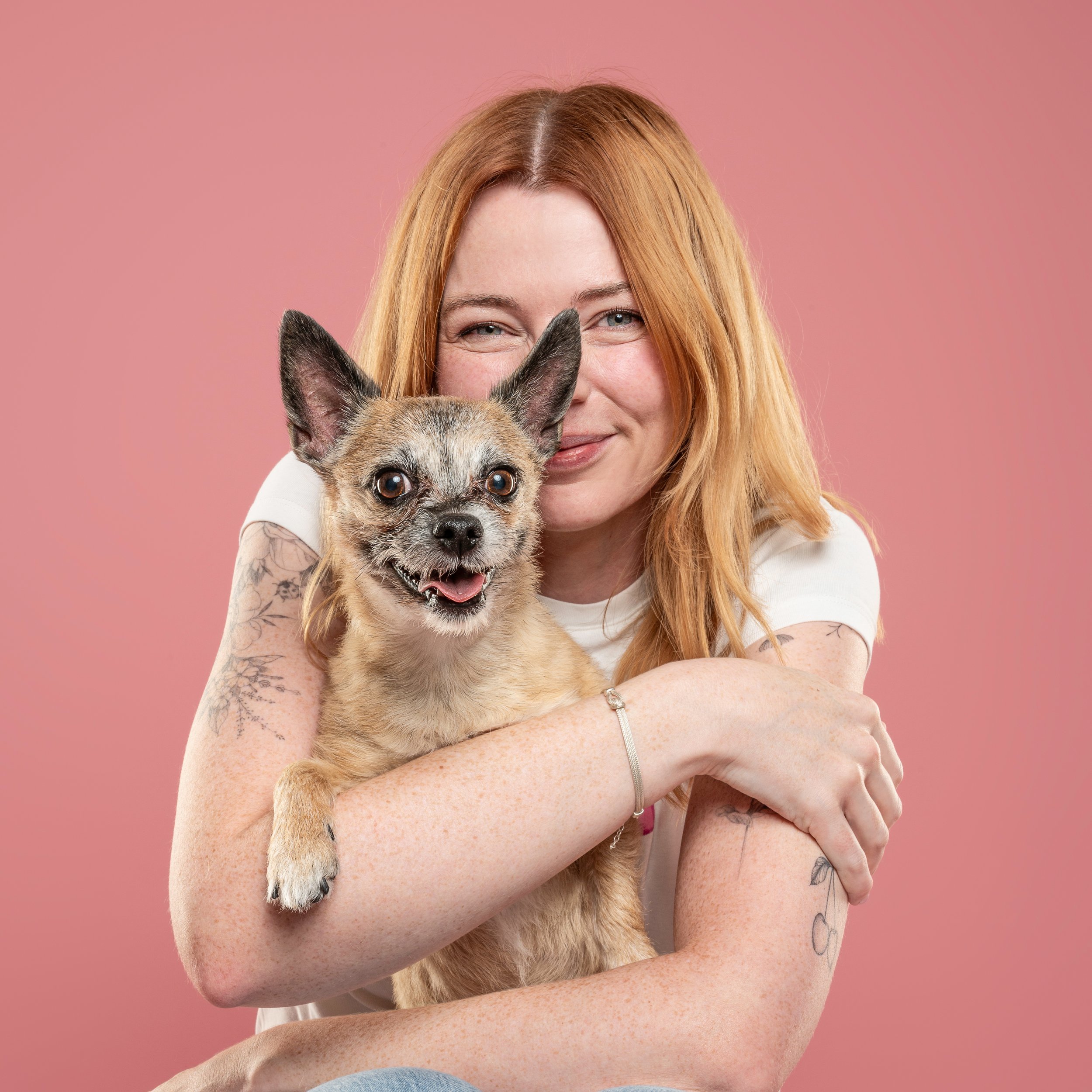 A woman with long red hair and tattoos is smiling while holding a small dog with a brindle coat and large ears in front of a pink background.