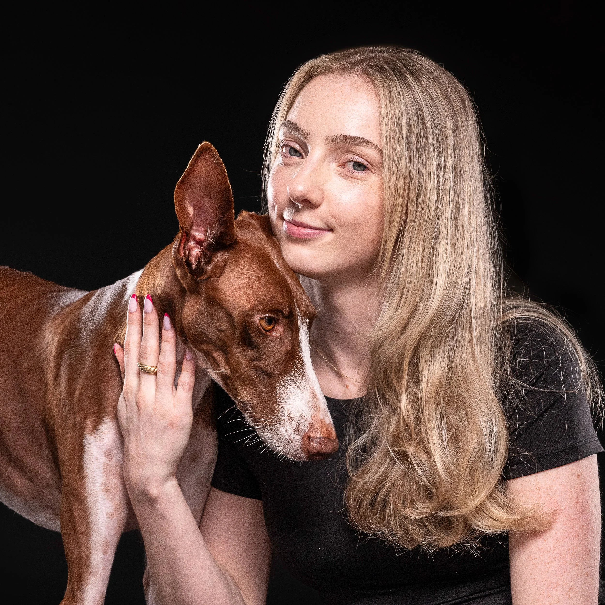 A young woman with long blonde hair and blue eyes, smiling softly, holding a brown and white dog with large ears against a black background.