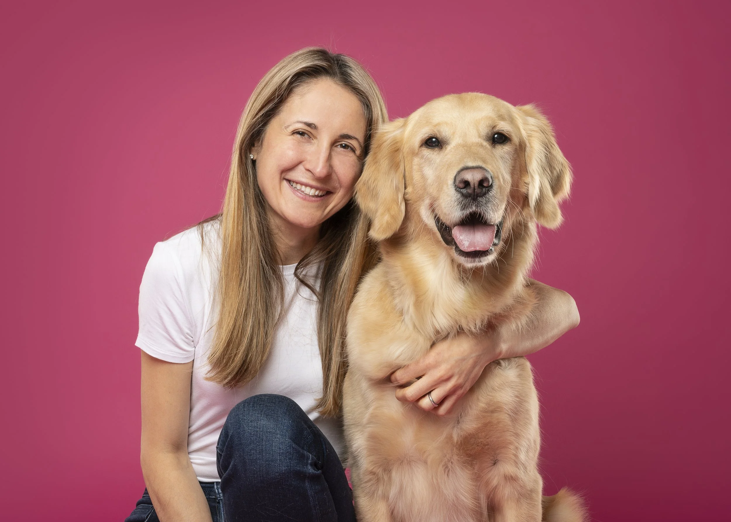A smiling woman with long blonde hair holding a golden retriever puppy against a pink background.