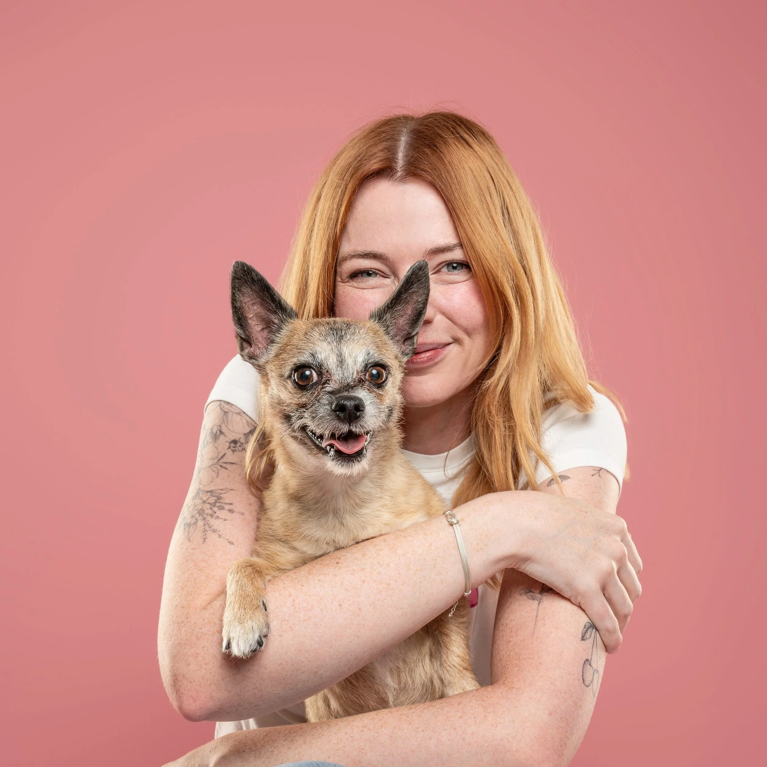 A woman with red hair holding a dog with a brindle coat and large ears, against a pink background.
