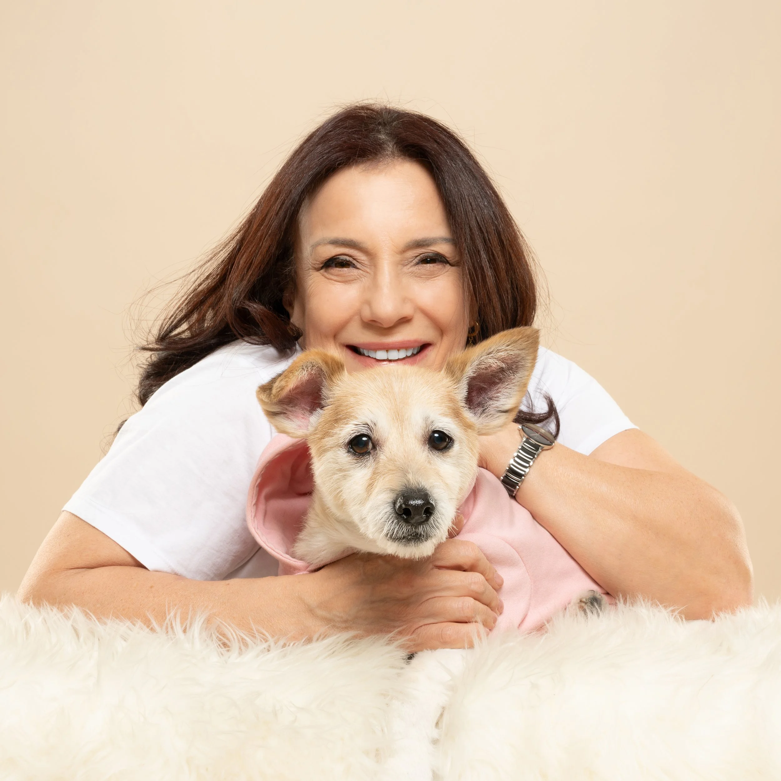 A woman with brown hair smiling while holding a light-colored puppy, with a beige wall background.
