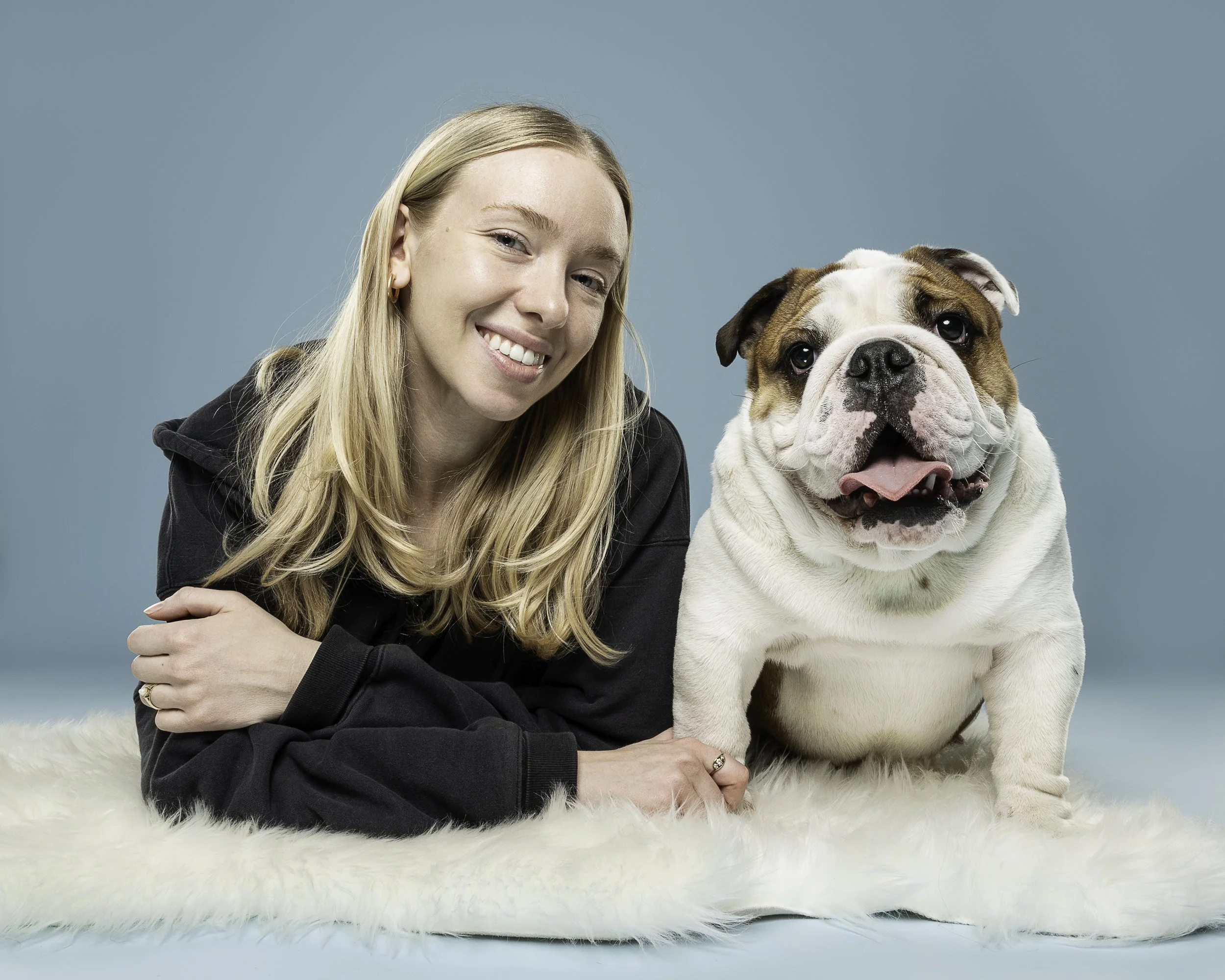 A young woman with long blonde hair, wearing a black hoodie, smiling and leaning on a large bulldog sitting on a white fur rug, against a solid light blue background.