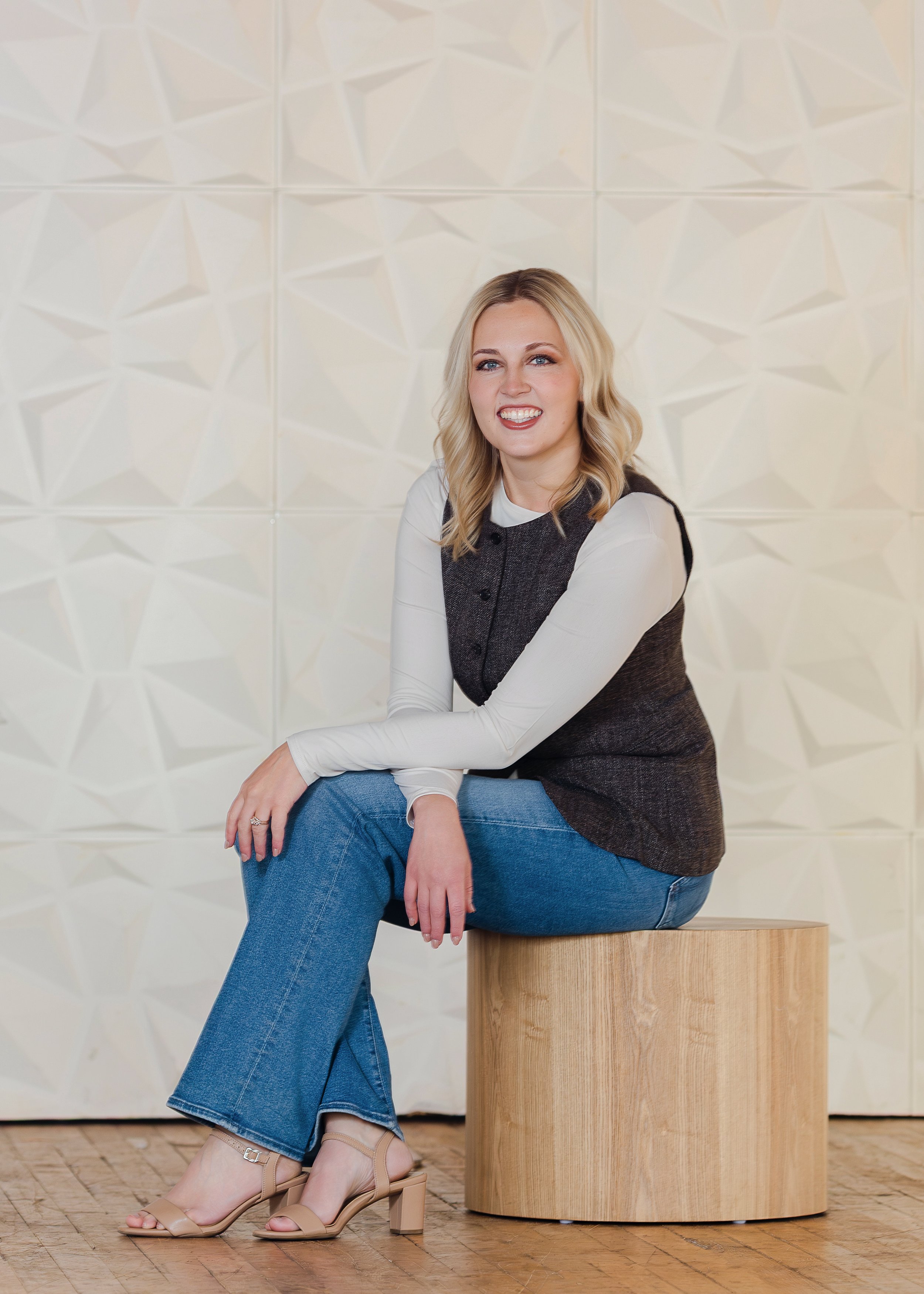 A woman with blonde hair, wearing a beige blazer over a white top, sitting on a white stool in front of a textured white wall.