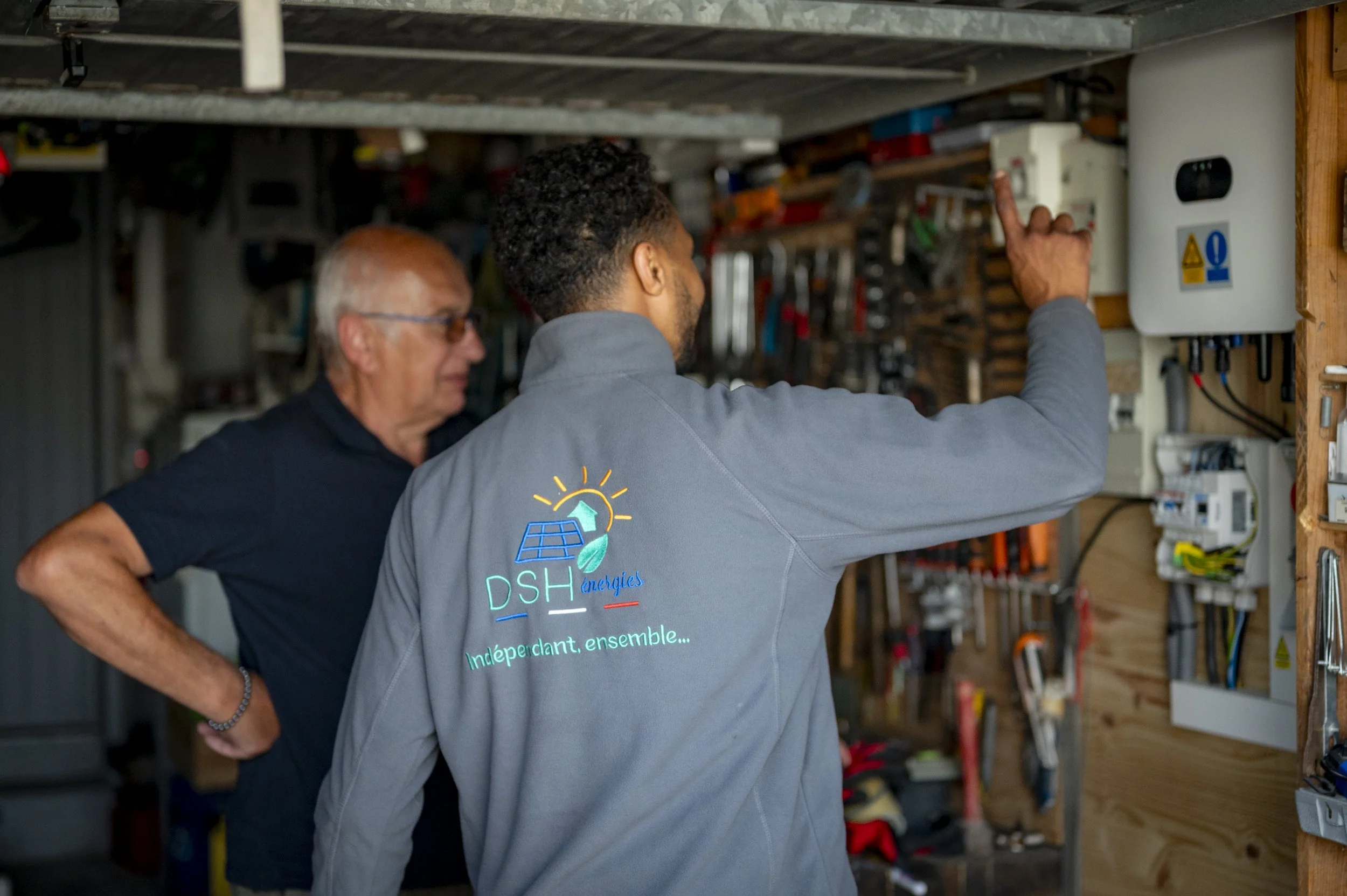 Deux hommes discutent dans un atelier, un d'eux porte une veste grise avec un logo d'énergie solaire et le texte 'DSH énergies' et 'Indépendant, ensemble'