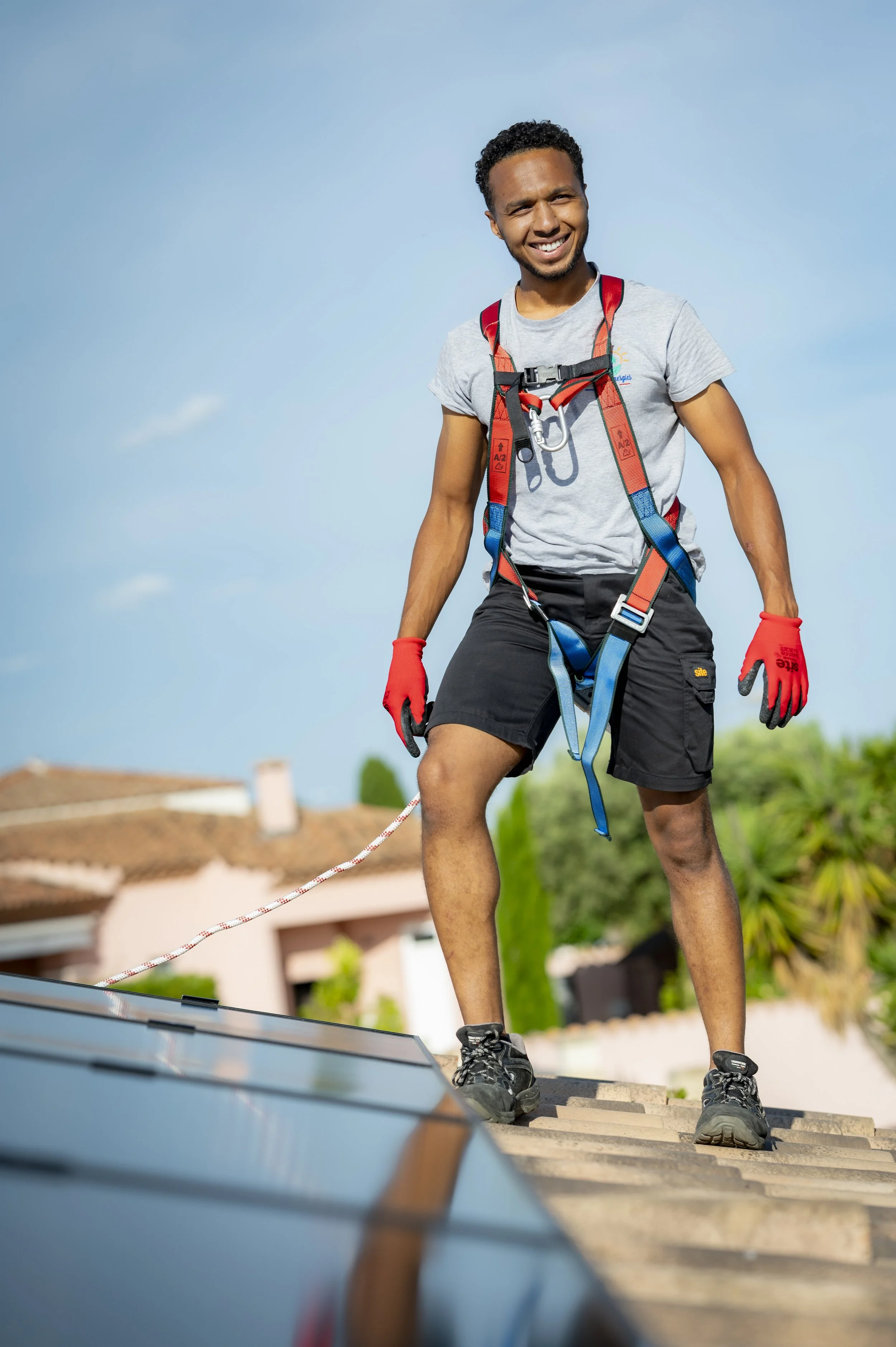 Homme en tenue d'escalade sur un toit, attaché en sécurité, souriant, avec un ciel bleu en arrière-plan.