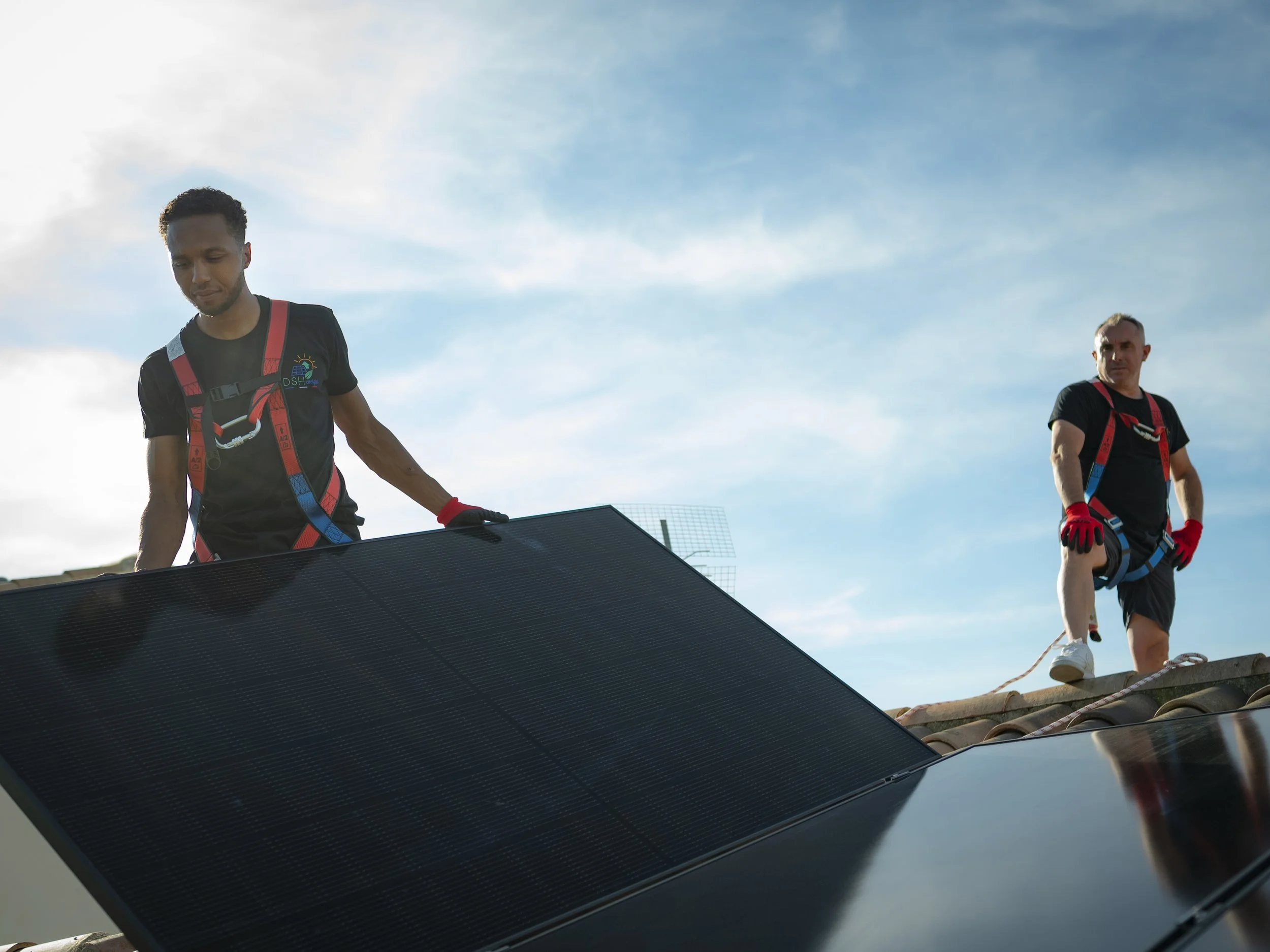 Deux hommes travaillent sur un toit, installant des panneaux solaires, portant des équipements de sécurité et des gants rouges, sous un ciel bleu.