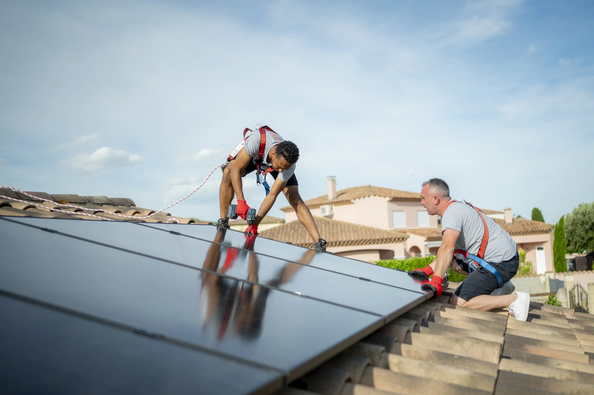 Deux hommes installent des panneaux solaires sur le toit d'une maison résidentielles, sous un ciel bleu avec quelques nuages.
